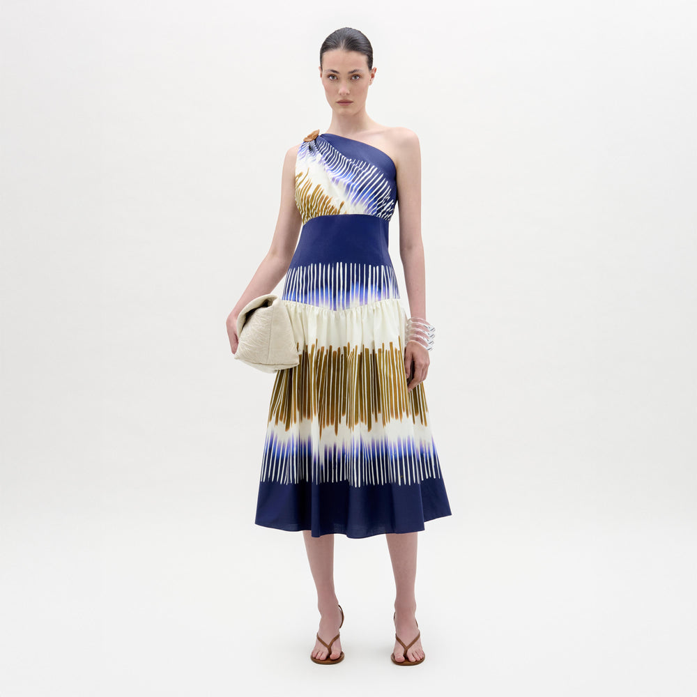 A woman stands against a plain background wearing the Catalina Dress, a one-shoulder blue and white patterned midi with an A-line silhouette, paired with a beige clutch, sandals, and stacked bracelets.