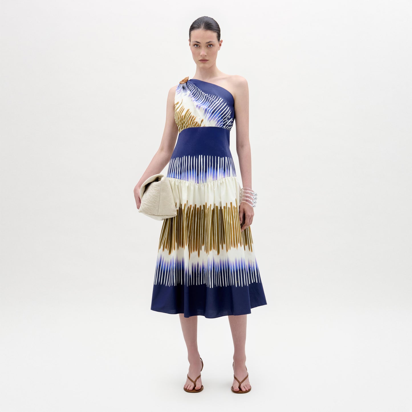 A woman stands against a plain background wearing the Catalina Dress, a one-shoulder blue and white patterned midi with an A-line silhouette, paired with a beige clutch, sandals, and stacked bracelets.