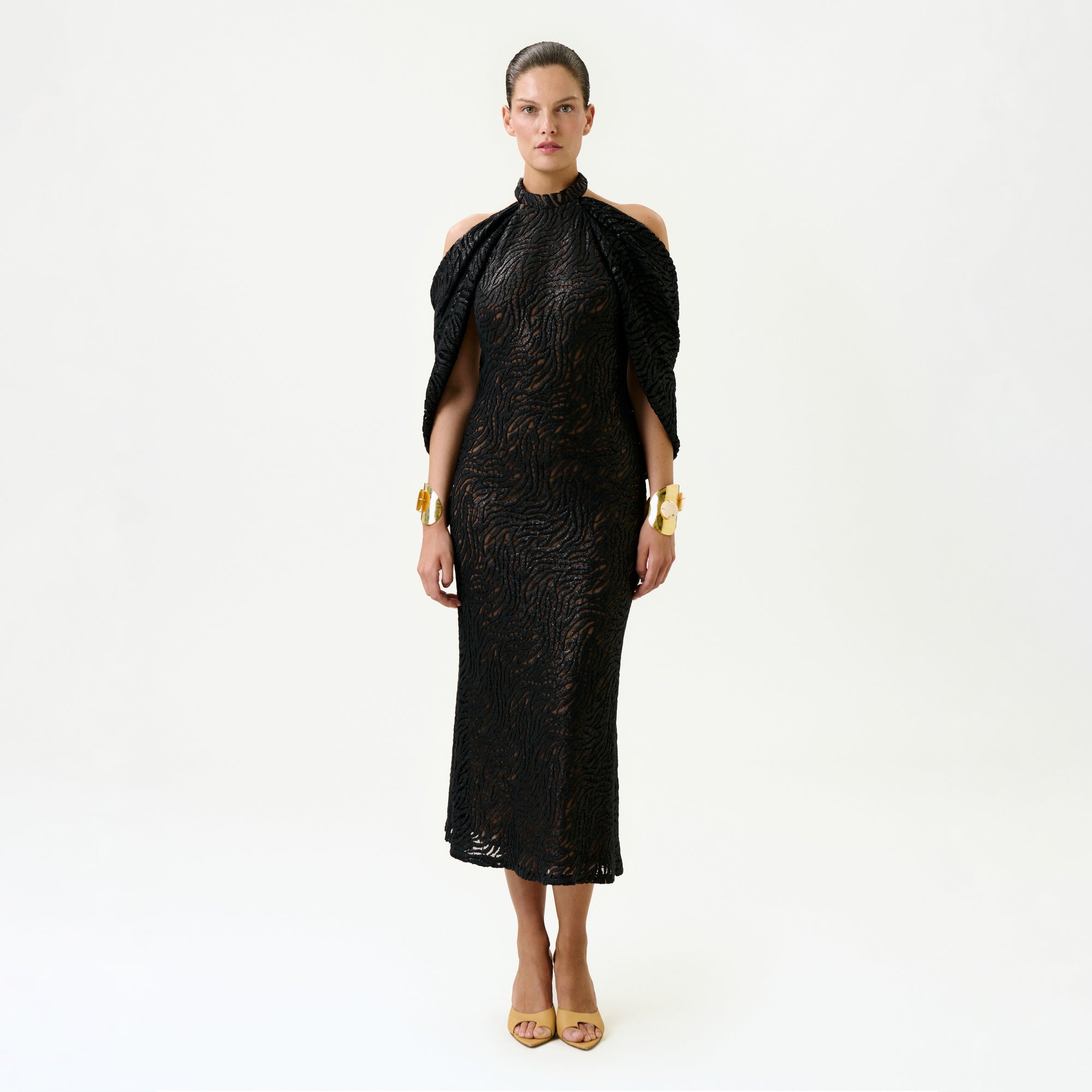 A woman stands facing forward against a plain white background, wearing the Neda Dress—a black, fitted, textured midi dress with cut-out shoulders—paired with gold bracelets and beige open-toe heels. Made in Colombia.
