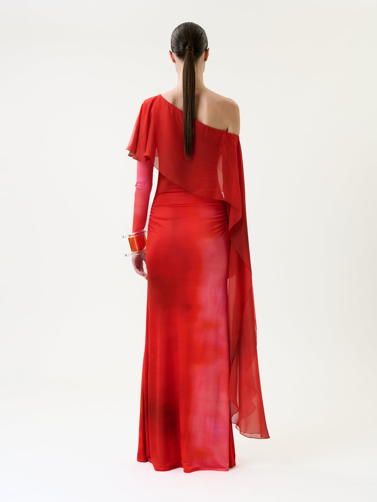 A person with long brown hair in a low ponytail wears the Maddy Dress, a one-shoulder, floor-length red gown featuring sheer draping and translucent red gloves, standing against a plain white background and facing away.