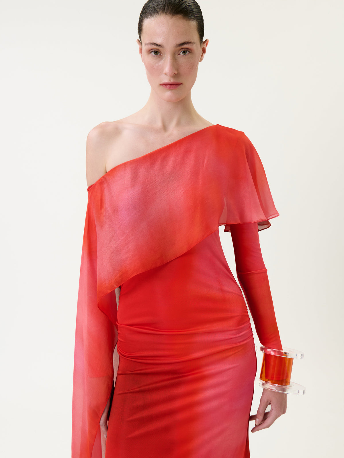 A woman wears the Maddy Dress, a sheer, asymmetrical red dress with one sleeve and one bare shoulder, standing against a plain light background. She accessorizes with a clear bracelet featuring a red accent on her left wrist.