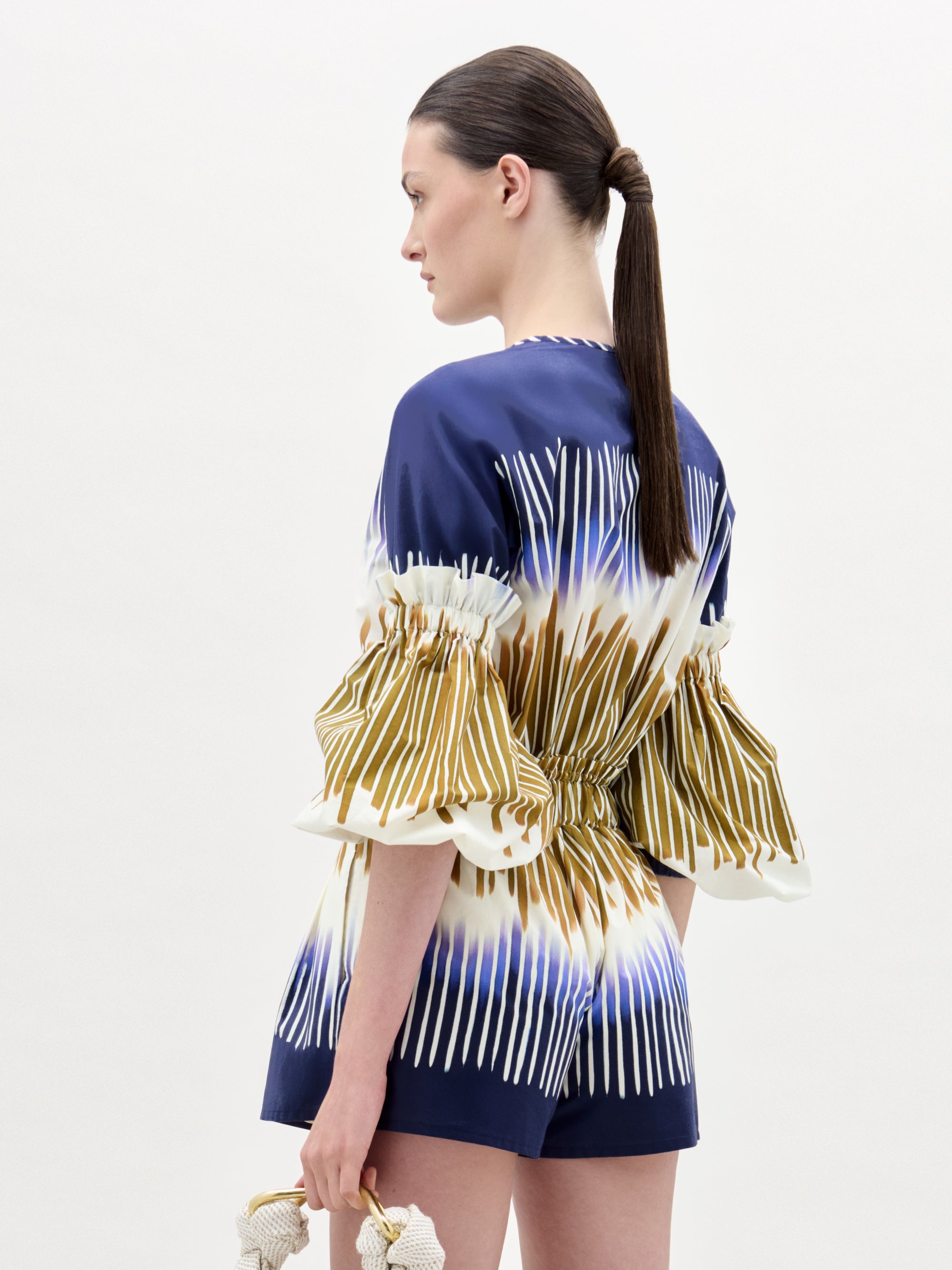 A woman with straight brown hair in a low ponytail wears the Catalina Dress, a blue and white patterned midi dress with gold stripes and puff sleeves. She holds a small textured clutch and faces away from the camera against a plain background.