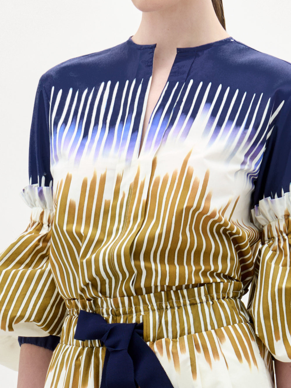 A woman stands against a plain background, wearing the Lucaya Blouse—an Italian fabric top in blue and white patterns with gold stripes, puffed sleeves, a belted waist, and elegant detailing.