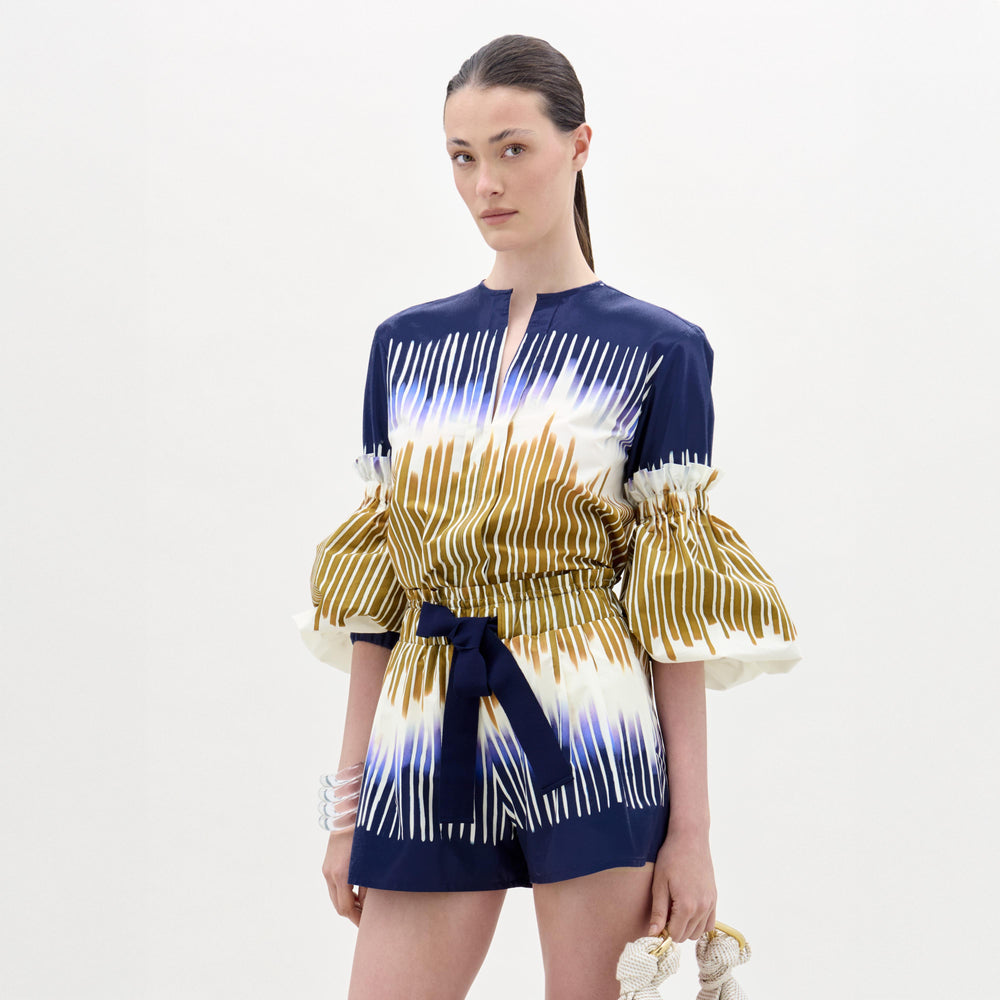 A woman stands against a plain background, wearing the Lucaya Blouse—an Italian fabric top in blue and white patterns with gold stripes, puffed sleeves, a belted waist, and elegant detailing.