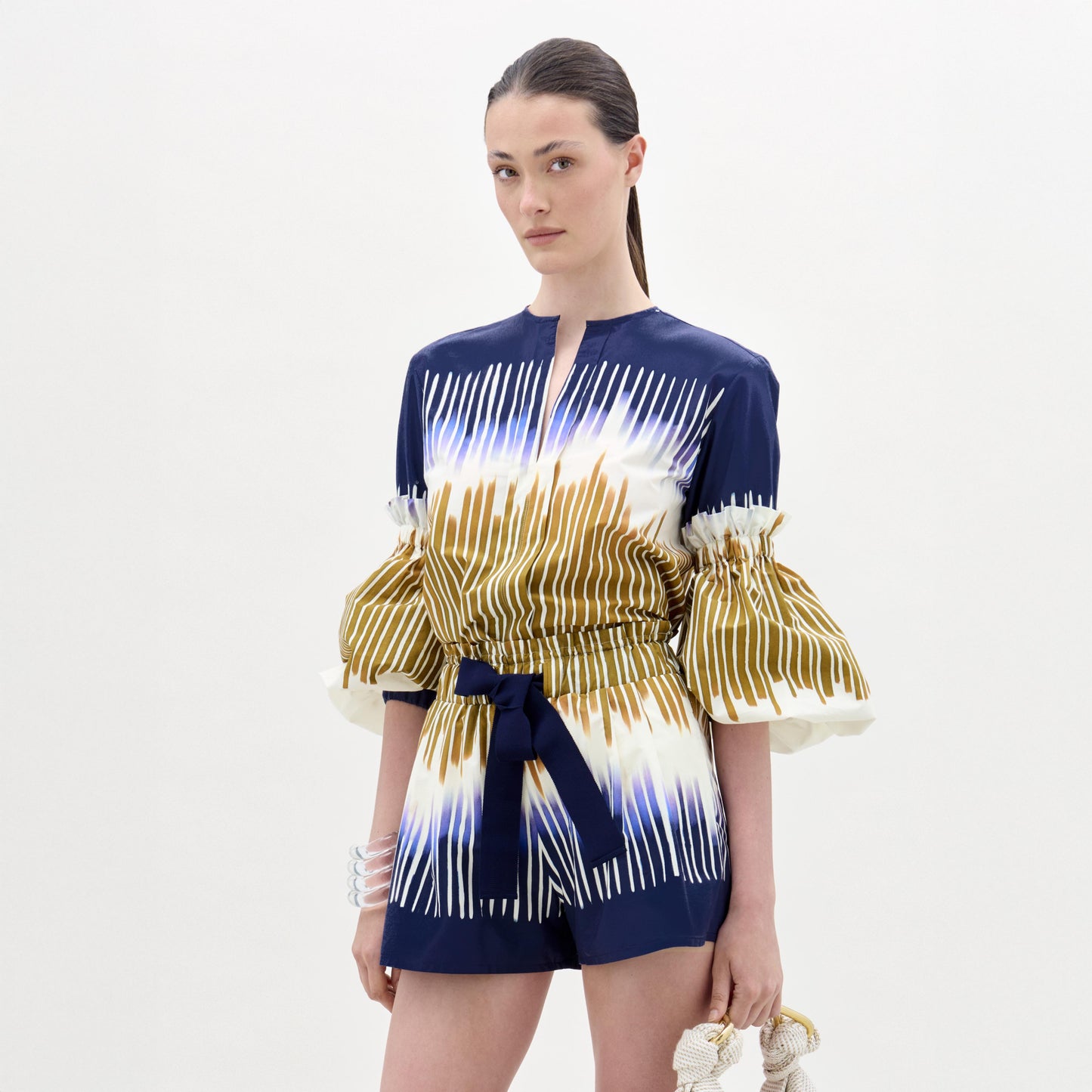 A woman stands against a plain background, wearing the Lucaya Blouse—an Italian fabric top in blue and white patterns with gold stripes, puffed sleeves, a belted waist, and elegant detailing.