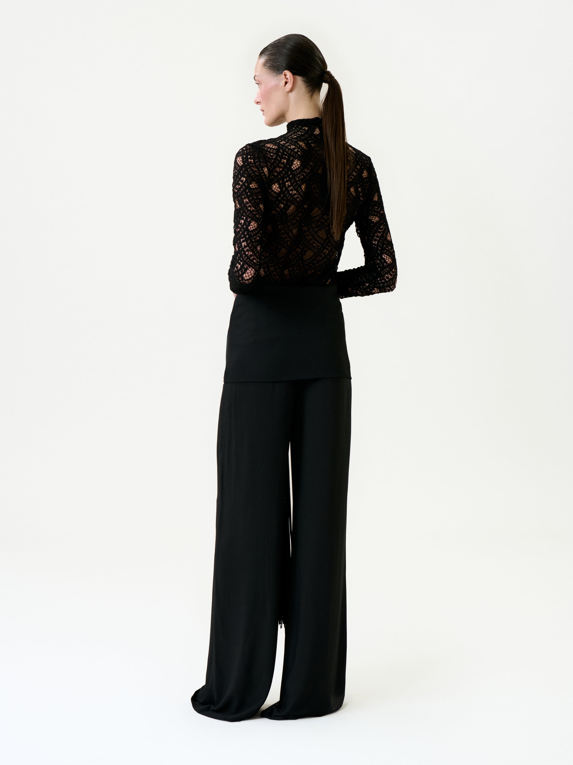 A woman faces away, styled in a black lace long-sleeve top and Kiele Pant—high-waisted, wide-leg, black pants made from premium Italian fabric. Her dark hair is tied in a low ponytail against a plain white background.