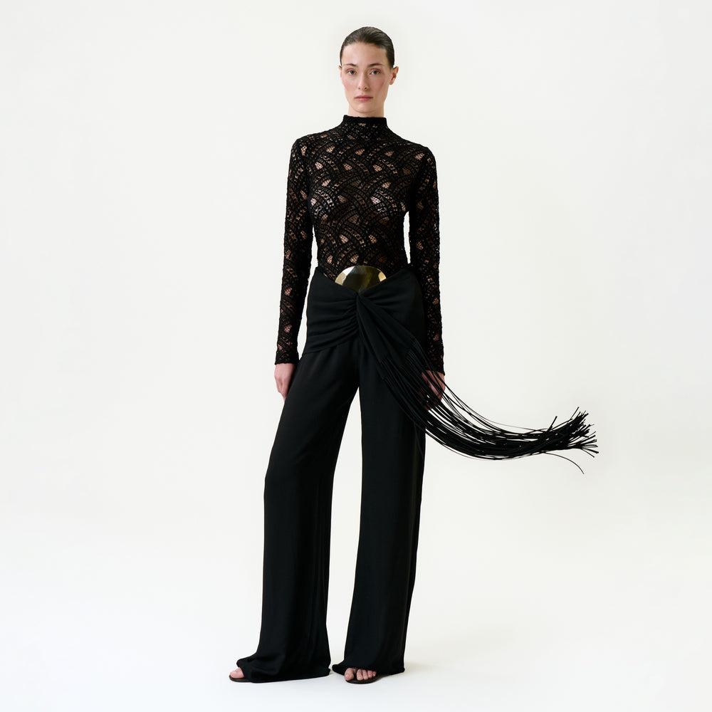 A woman stands against a plain white background wearing the Kiele Pant—high-waisted black pants with a large metallic belt and draped sash of long, flowing fringes, crafted from luxurious Italian fabric.