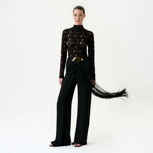 A woman stands against a plain white background wearing the Kiele Pant—high-waisted black pants with a large metallic belt and draped sash of long, flowing fringes, crafted from luxurious Italian fabric.