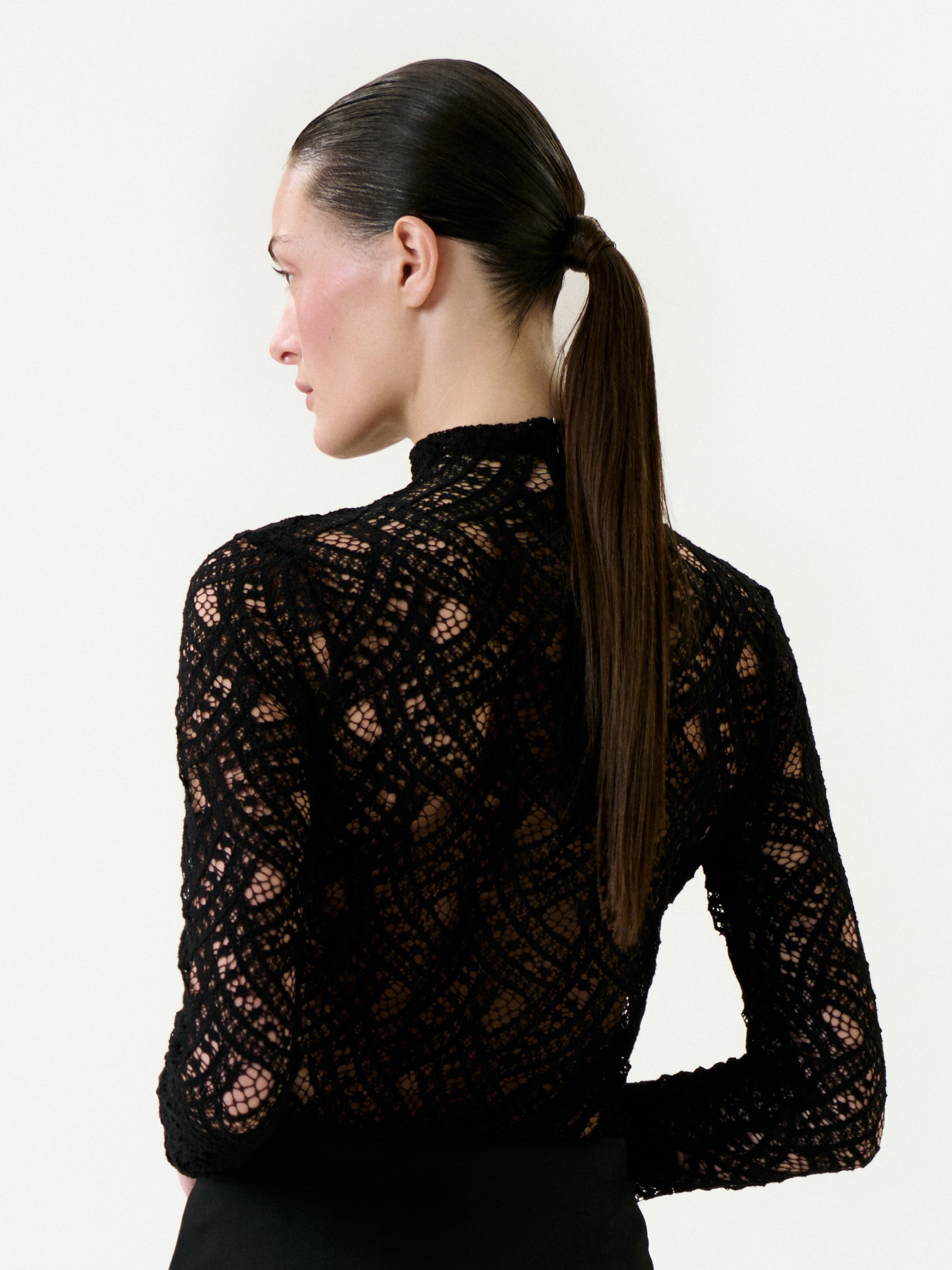 A woman with straight brown hair in a low ponytail is seen from the side and back, wearing the Liz Blouse—a high-neck, long-sleeved black lace top with an intricate Italian pattern. The background is plain white.