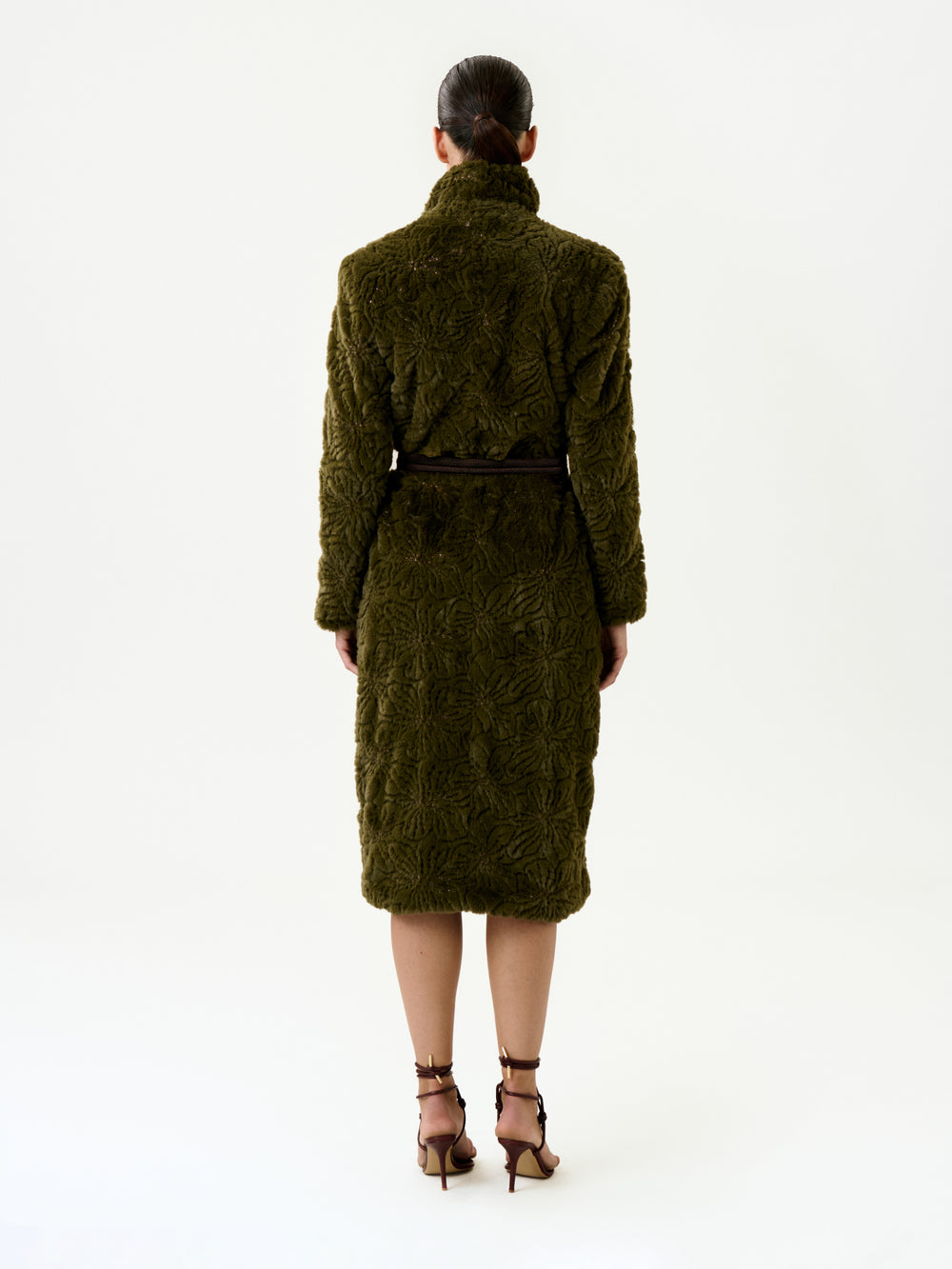 A woman stands facing away against a plain white background, wearing the Emerson Trench—a knee-length, textured olive green coat crafted from luxurious Italian fabric in an oversized fit—cinched at the waist with a black belt and paired with brown heels.