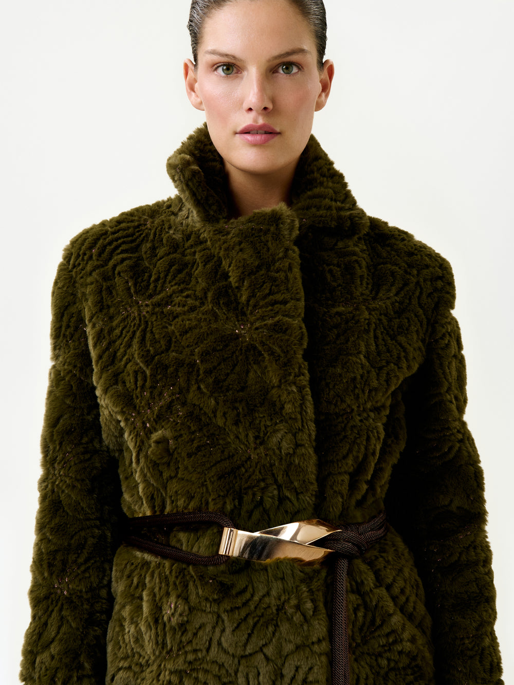 A woman wearing the Emerson Trench, a textured dark green faux fur coat with an oversized fit and a shiny metallic belt, stands against a plain light background.