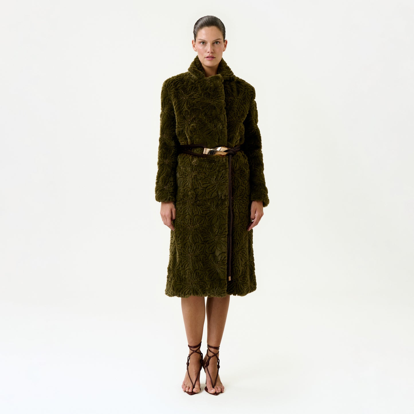 A woman stands facing forward against a plain white background, wearing the Emerson Trench—a long, textured dark green fur trench coat crafted from luxurious Italian fabric, featuring a wide collar and a thin brown belt—paired with strappy brown high-heeled sandals.