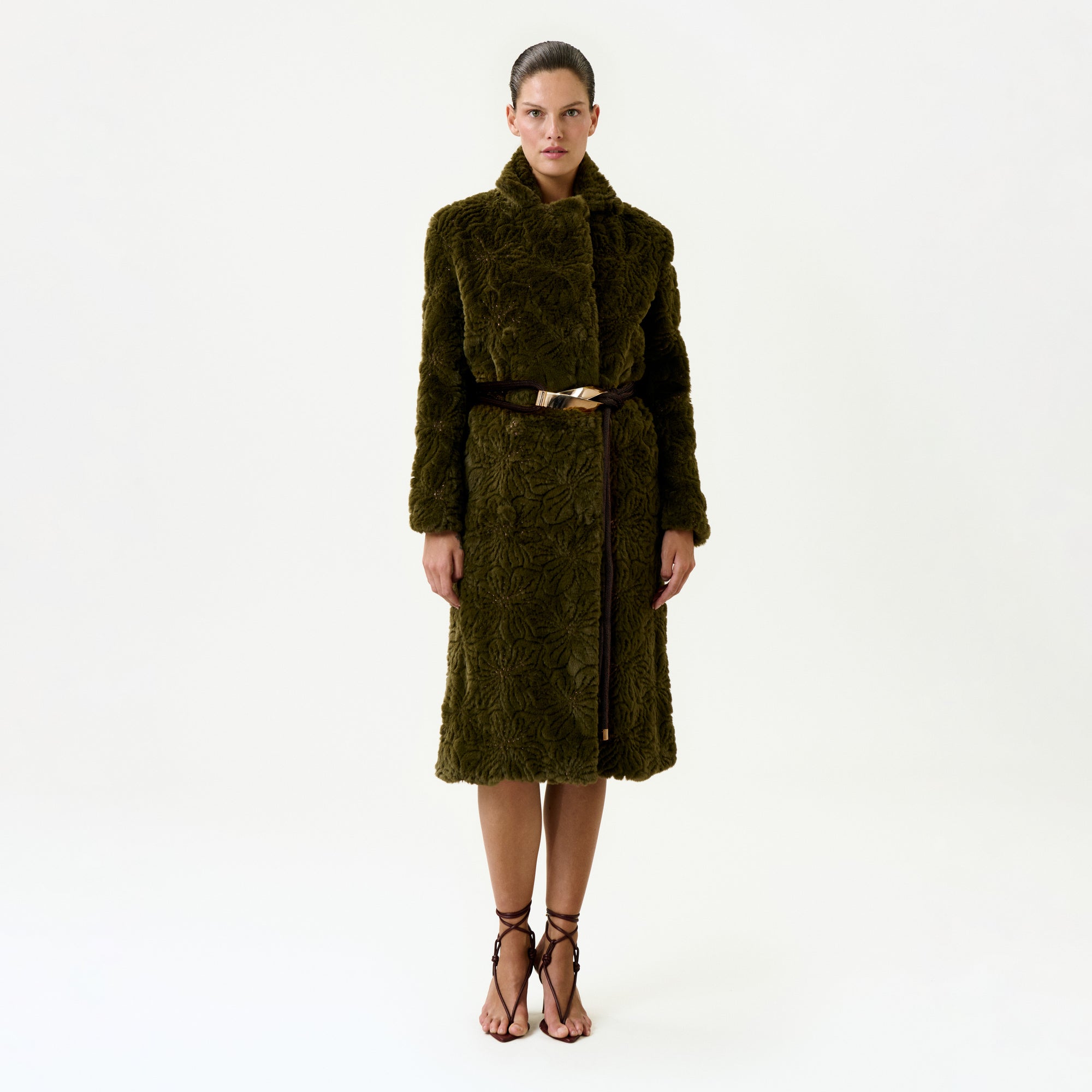 A woman stands facing forward against a plain white background, wearing the Emerson Trench—a long, textured dark green fur trench coat crafted from luxurious Italian fabric, featuring a wide collar and a thin brown belt—paired with strappy brown high-heeled sandals.