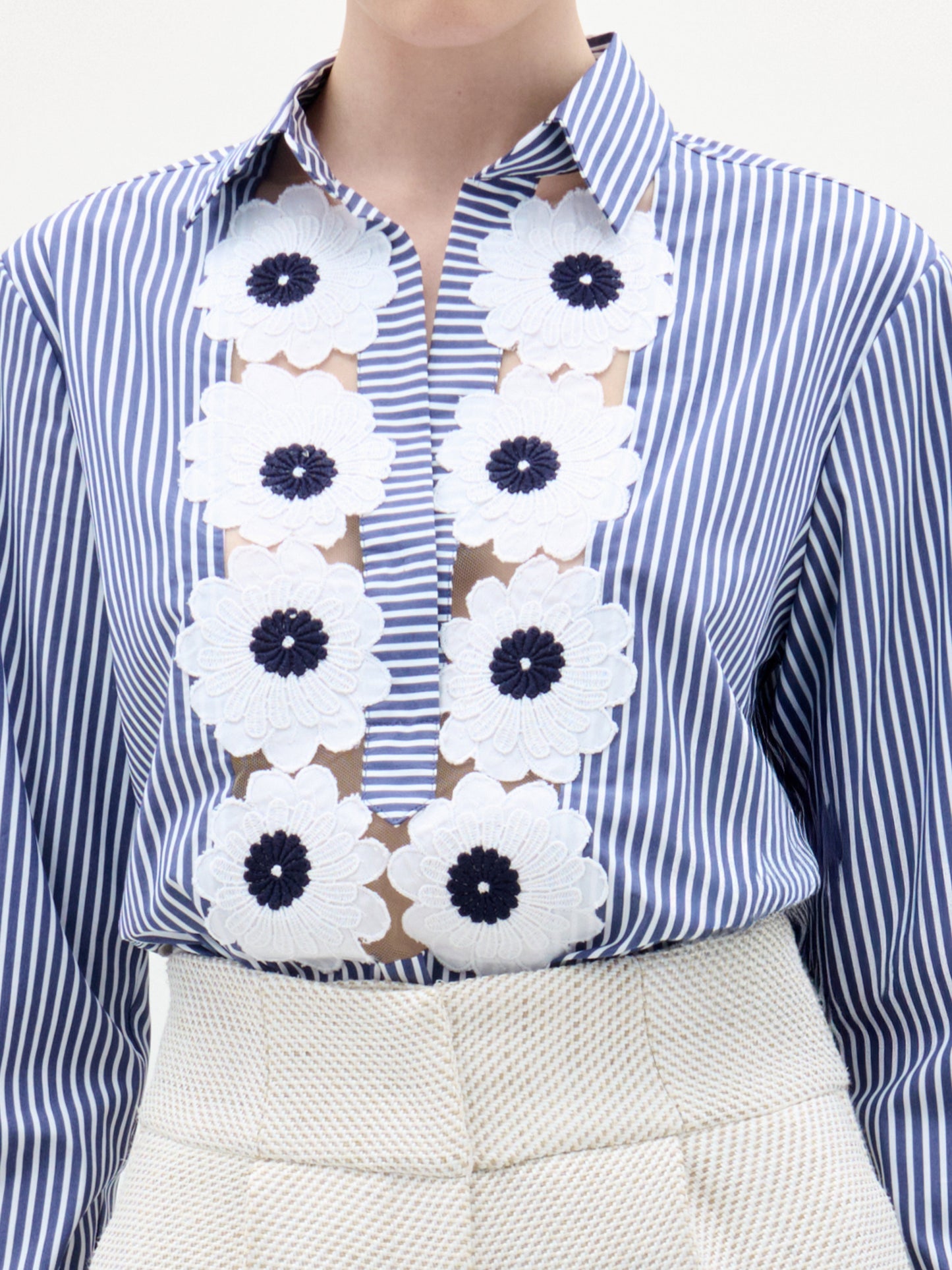 A woman stands against a plain background wearing the Karole Blouse, a blue and white striped button-down with floral embroidery made from Italian fabric, paired with high-waisted cream pants.