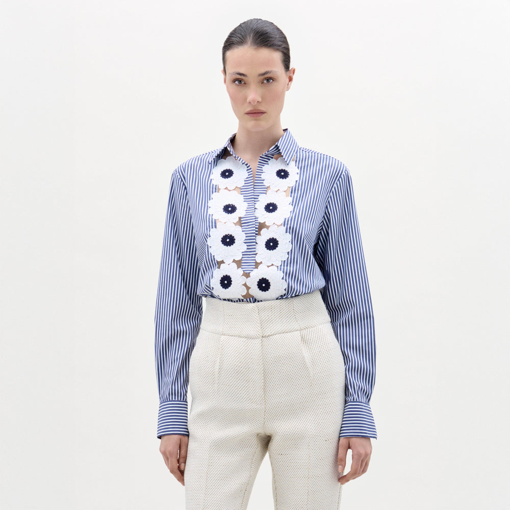 A woman stands against a plain background wearing the Karole Blouse, a blue and white striped button-down with floral embroidery made from Italian fabric, paired with high-waisted cream pants.