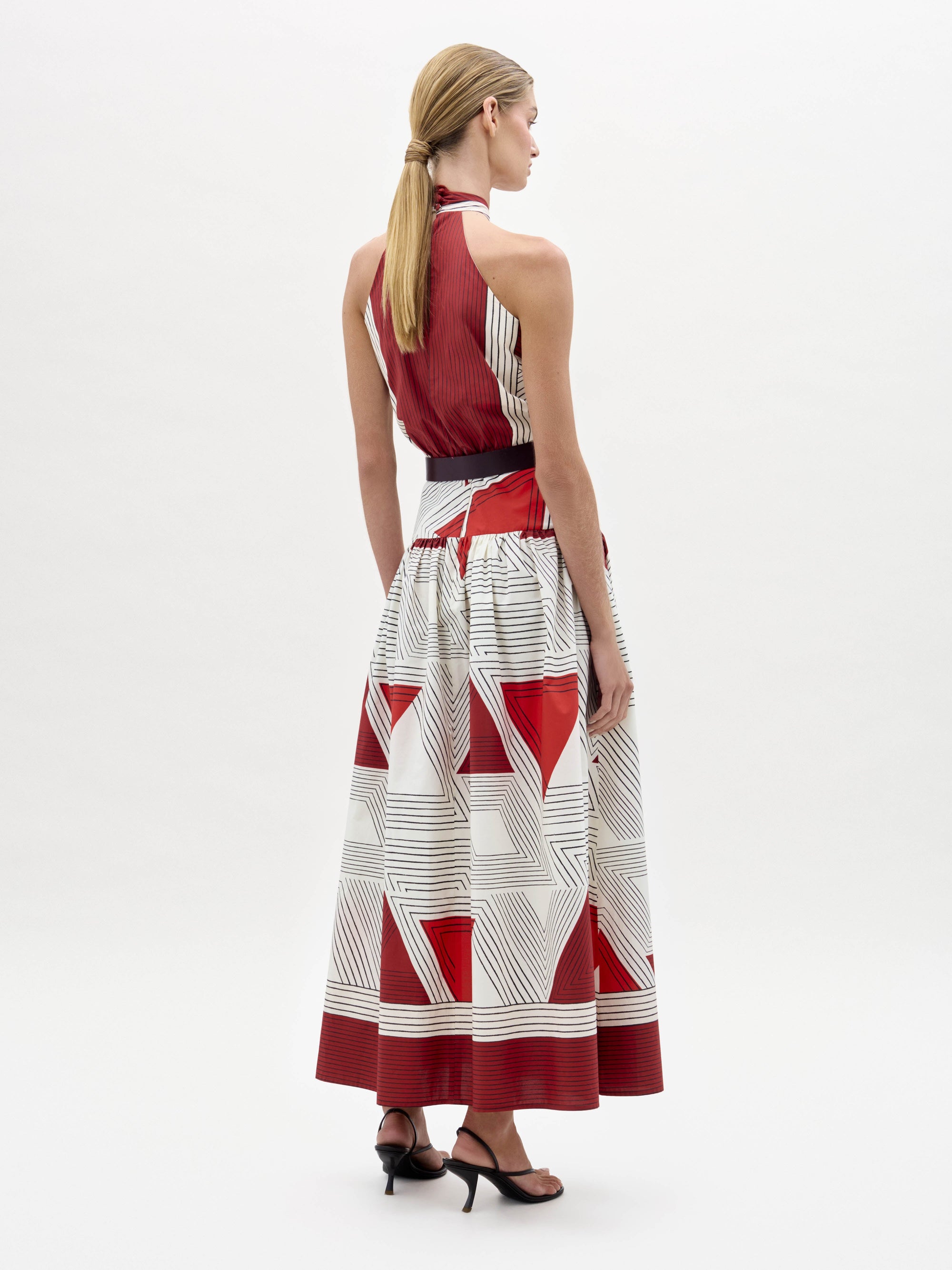 A woman wearing the Lana Dress—a halter-neck, floor-length dress with red and white geometric patterns—paired with a wide belt and black sandals, stands against a plain white background.