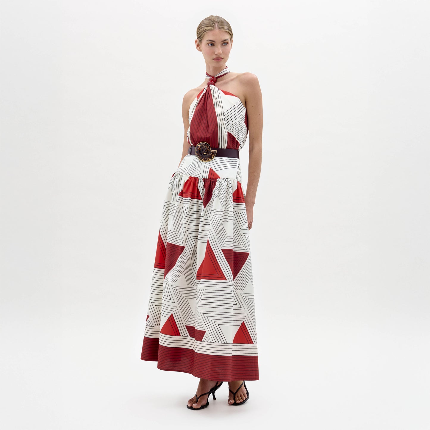 A woman wearing the Lana Dress—a halter-neck, floor-length dress with red and white geometric patterns—paired with a wide belt and black sandals, stands against a plain white background.