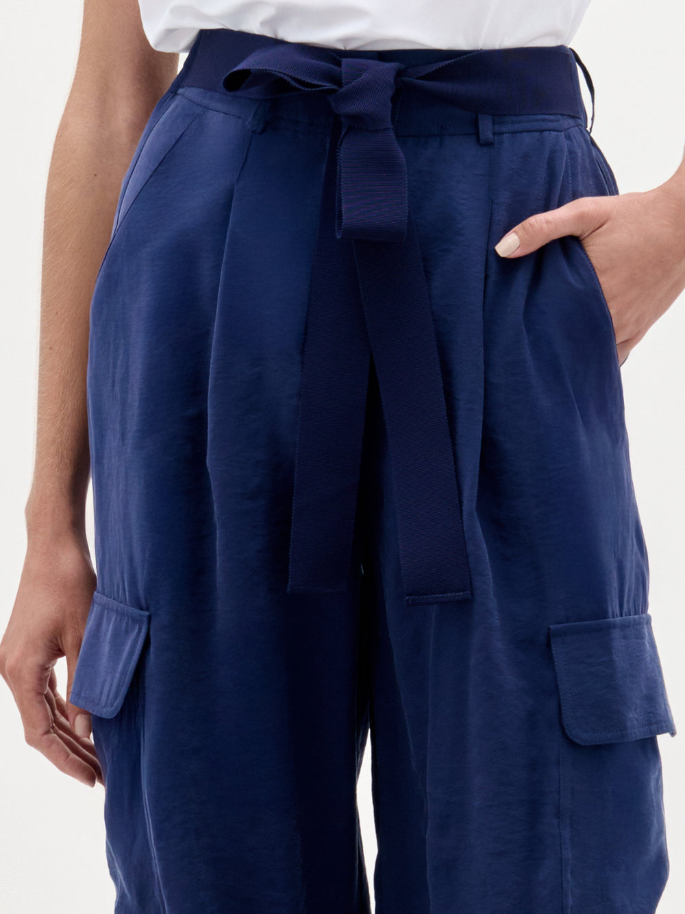 A woman stands against a plain background wearing a white one-shoulder top, brown sandals, and the Darnell Pant—navy high-waisted cargo pants made from Italian fabric.