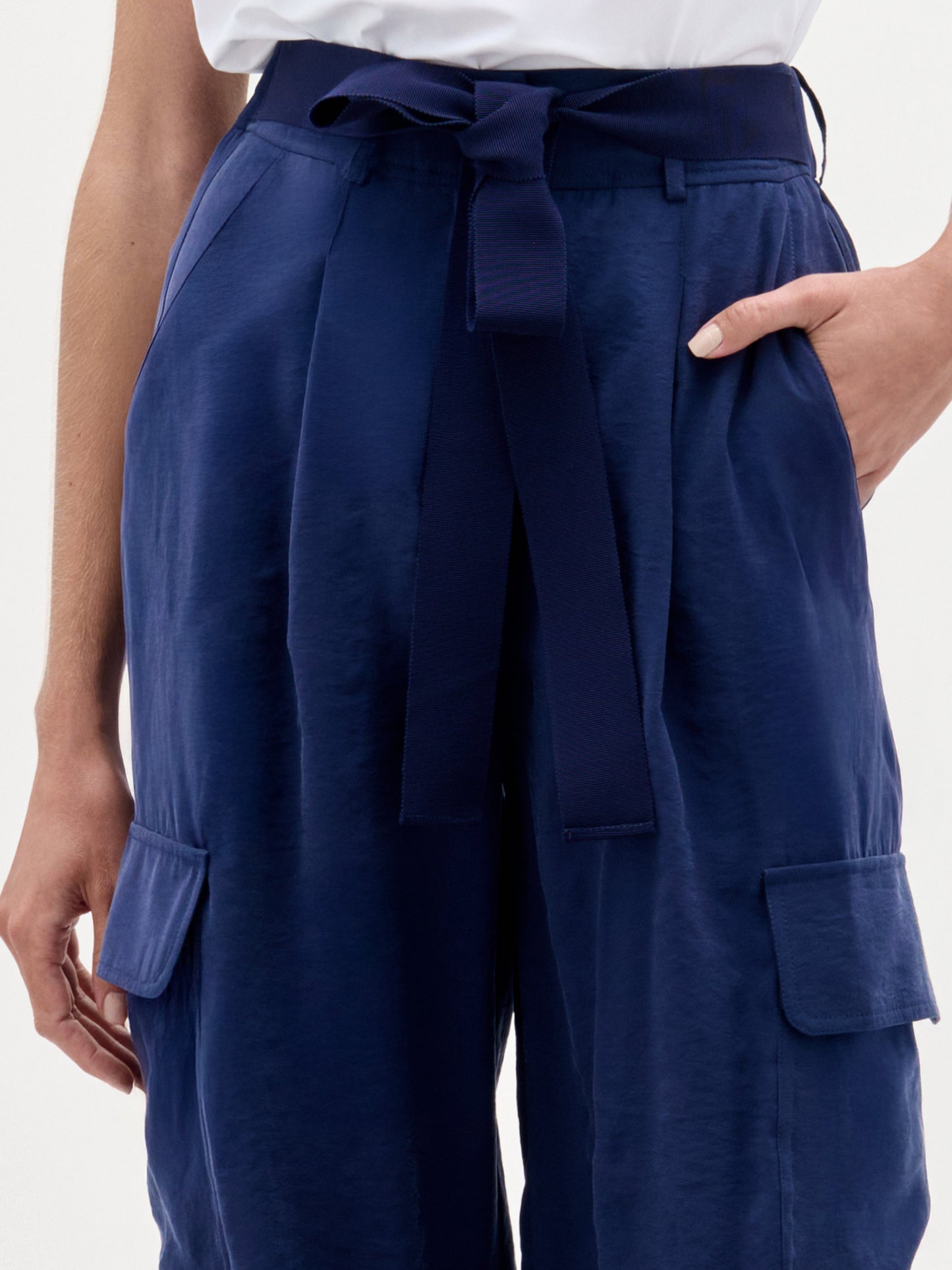 A woman stands against a plain background wearing a white one-shoulder top, brown sandals, and the Darnell Pant—navy high-waisted cargo pants made from Italian fabric.
