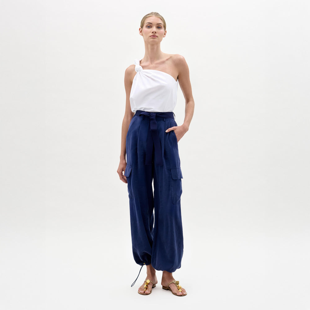 A woman stands against a plain background wearing a white one-shoulder top, brown sandals, and the Darnell Pant—navy high-waisted cargo pants made from Italian fabric.