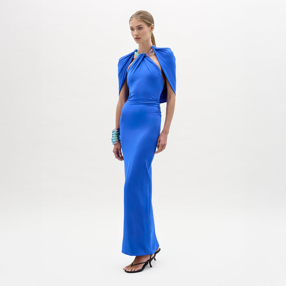 Woman in the Odele Dress, a fitted, floor-length bright blue maxi with cape-like detail and halter neckline, stands against a white background wearing heeled sandals and matching blue bracelets.