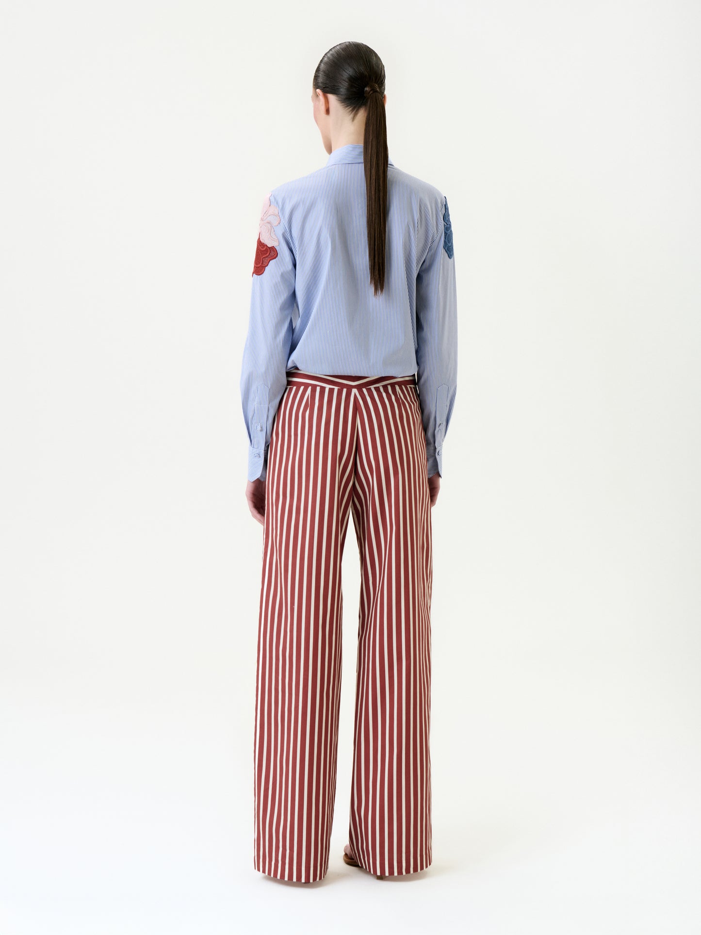 A person with long dark hair in a low ponytail faces away, wearing a light blue shirt with embroidered patches and the Ulla Pant—high-waisted, wide-leg red-and-white vertical striped pants made from 100% cotton.