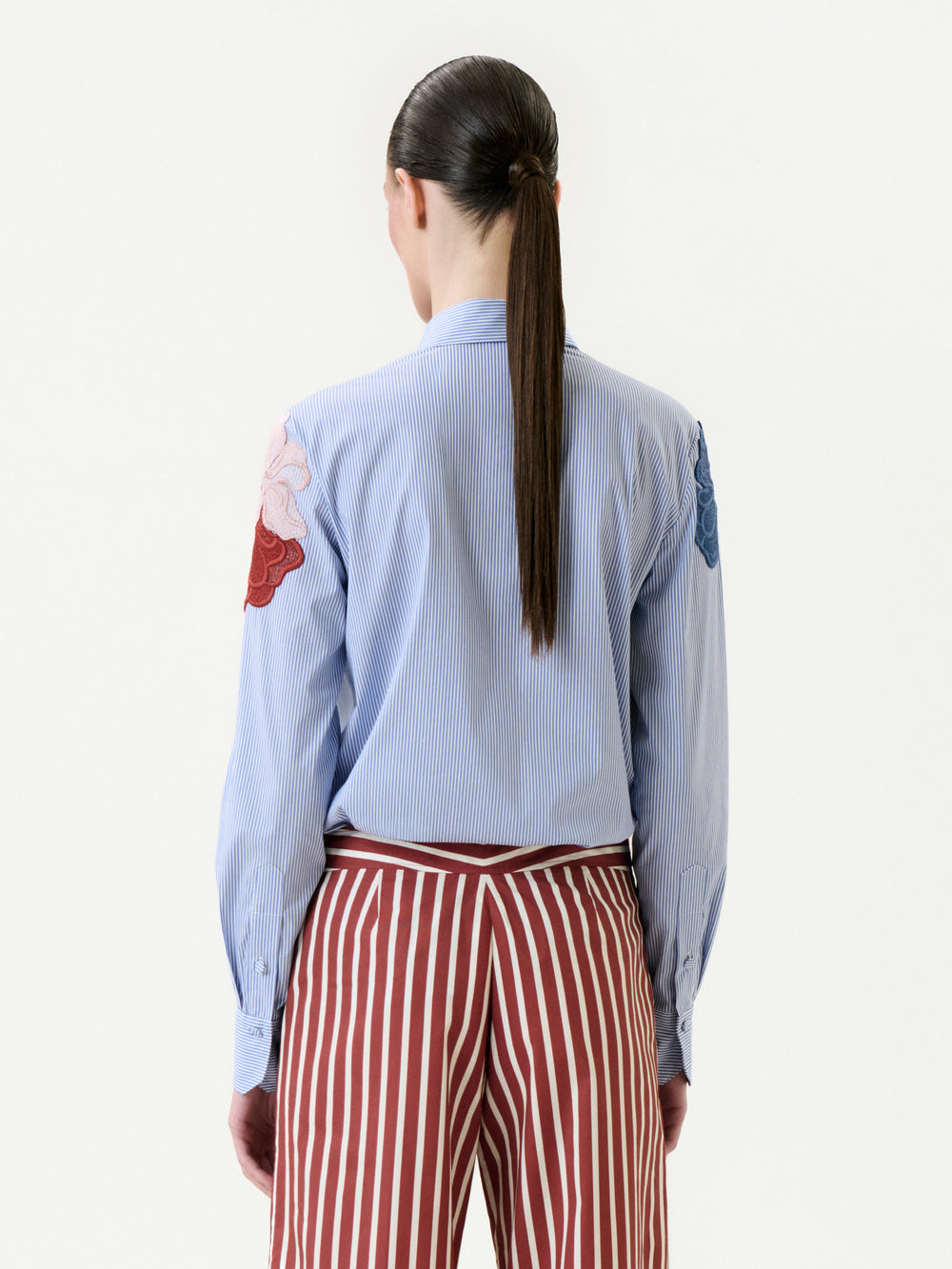 A person with long brown hair in a low ponytail is seen from behind, wearing the Warda Shirt—blue and white Italian fabric with floral patch sleeves—and red-and-white vertical striped pants.