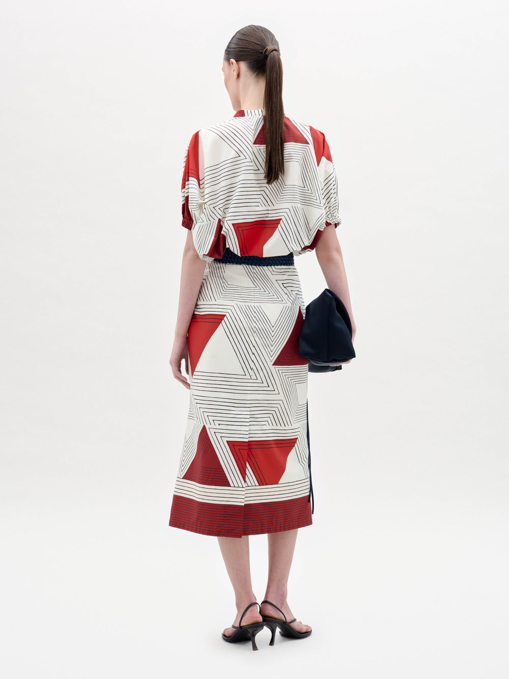 A woman stands against a plain background wearing the Atira Skirt paired with black sandals, a wide belt with a gold buckle, and holding a black bag at her side.