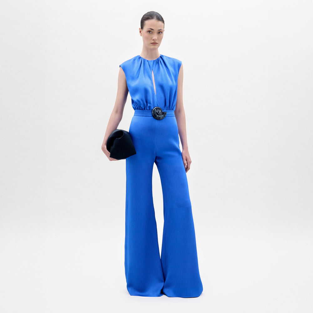 A woman stands against a plain background wearing the Noel Jumpsuit, a sleeveless bright blue wide-leg design with a gathered waist made from Italian textile, holding a black clutch.