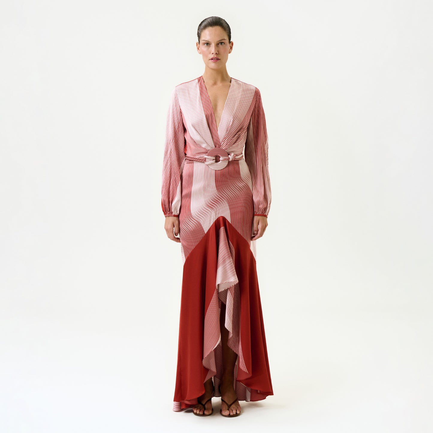 A woman stands facing forward in the Mairet Dress—a long, flowing silk maxi with pink and red geometric patterns, deep V-neck, long sleeves, wide belted waist with circular buckle, layered skirt, and hair pulled back.