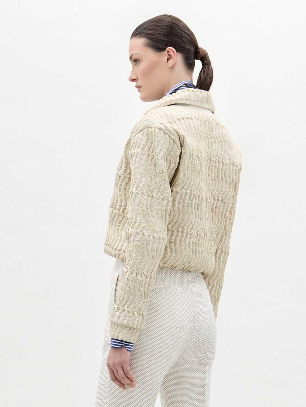 A woman with dark hair tied back models the Jaiden Jacket in textured cream Italian fabric over a blue-and-white striped shirt and cream pants, standing against a plain light background, facing away and slightly to the side.