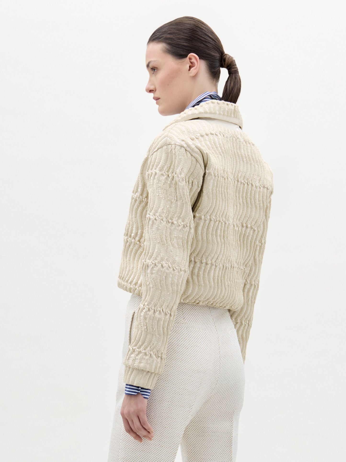 A woman with dark hair tied back models the Jaiden Jacket in textured cream Italian fabric over a blue-and-white striped shirt and cream pants, standing against a plain light background, facing away and slightly to the side.