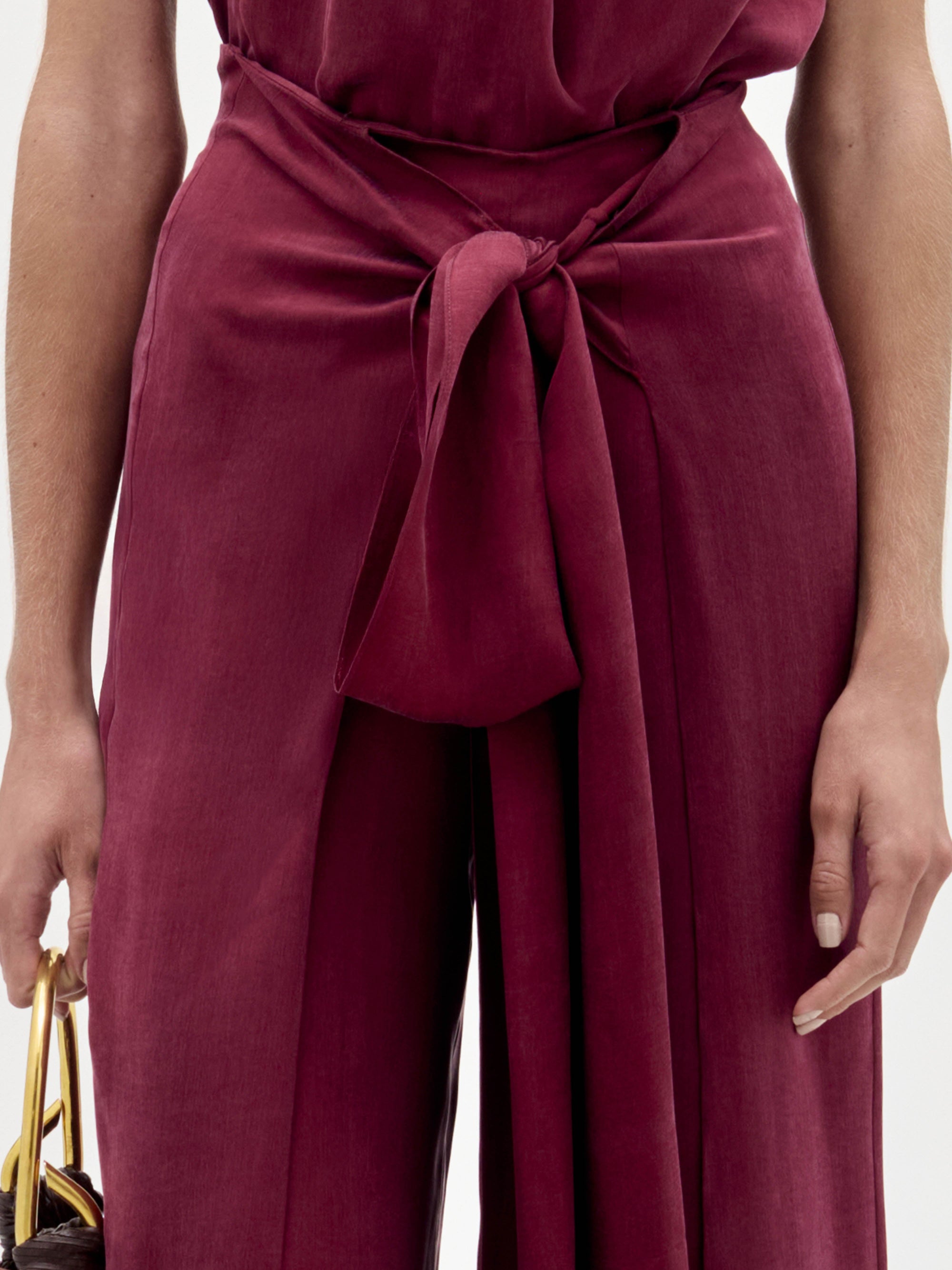 A woman stands against a white background wearing the Nuria Pant—a sleeveless maroon halter-neck jumpsuit with a wide waist tie—while holding a dark fringed handbag.