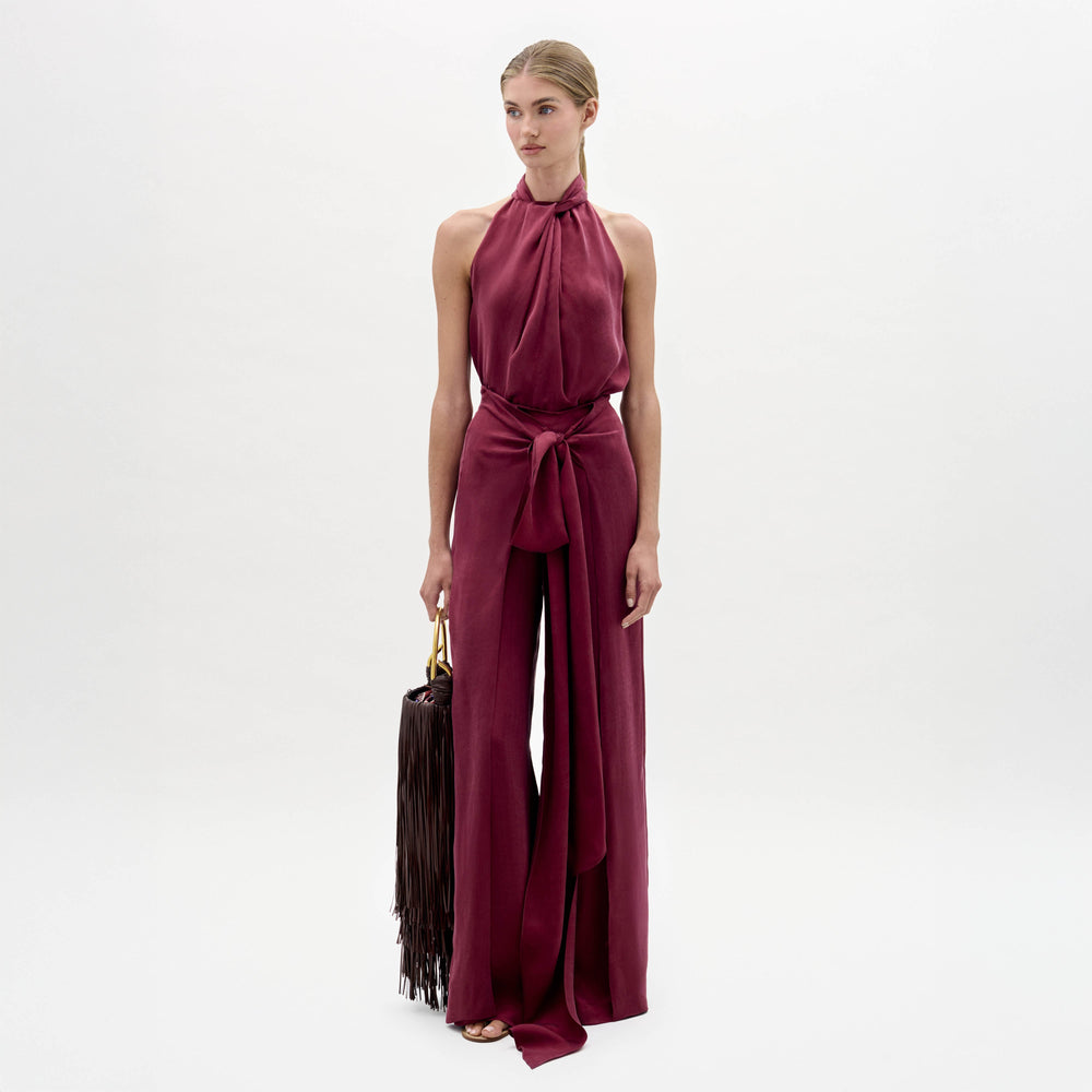 A woman stands against a white background wearing the Nuria Pant—a sleeveless maroon halter-neck jumpsuit with a wide waist tie—while holding a dark fringed handbag.