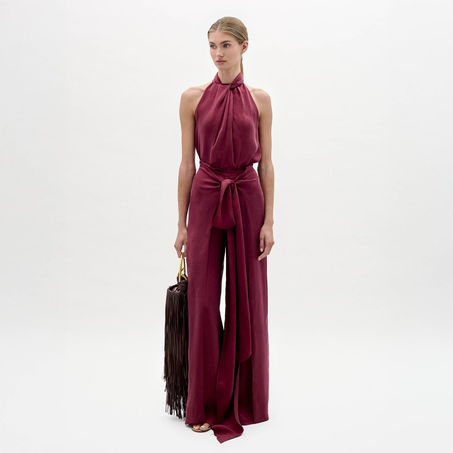 A woman stands against a white background wearing the Nuria Pant—a sleeveless maroon halter-neck jumpsuit with a wide waist tie—while holding a dark fringed handbag.