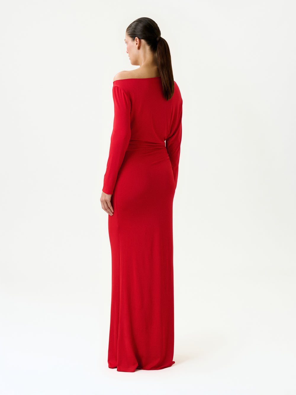 A woman with a ponytail models the Roxy Dress, a long-sleeved red off-the-shoulder maxi made in Colombia. She's standing with her back to the camera against a plain white background.