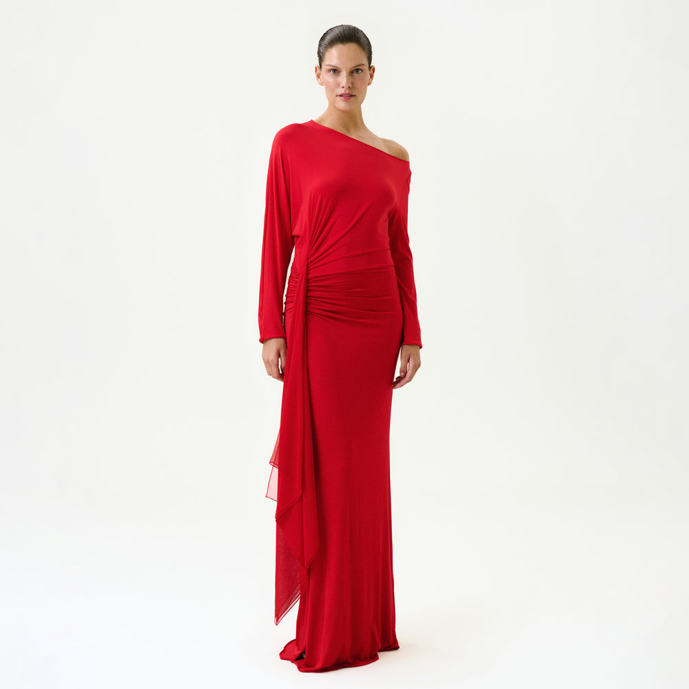 A woman with her hair pulled back poses confidently against a white background wearing the Roxy Dress, a long, fitted red maxi dress made in Colombia featuring a one-shoulder design, long sleeves, side draping, and an asymmetrical hem.
