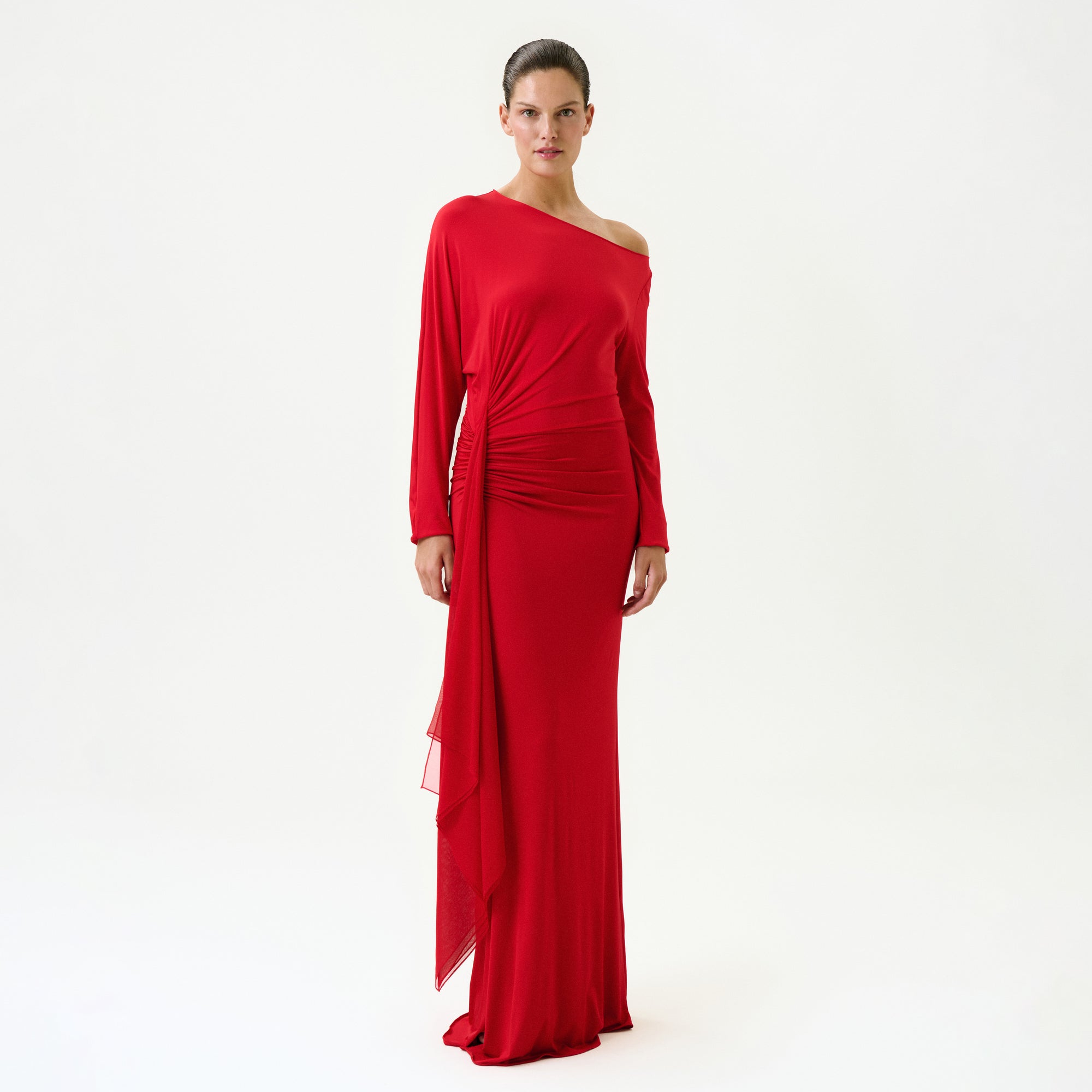 A woman with her hair pulled back poses confidently against a white background wearing the Roxy Dress, a long, fitted red maxi dress made in Colombia featuring a one-shoulder design, long sleeves, side draping, and an asymmetrical hem.