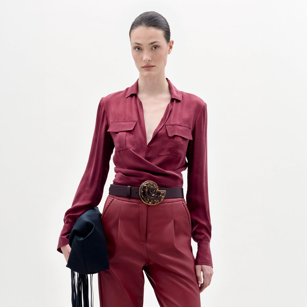 A woman stands against a plain background wearing the Caserta Blouse in deep red, matching pants, a large decorative belt, and holding a black clutch bag.