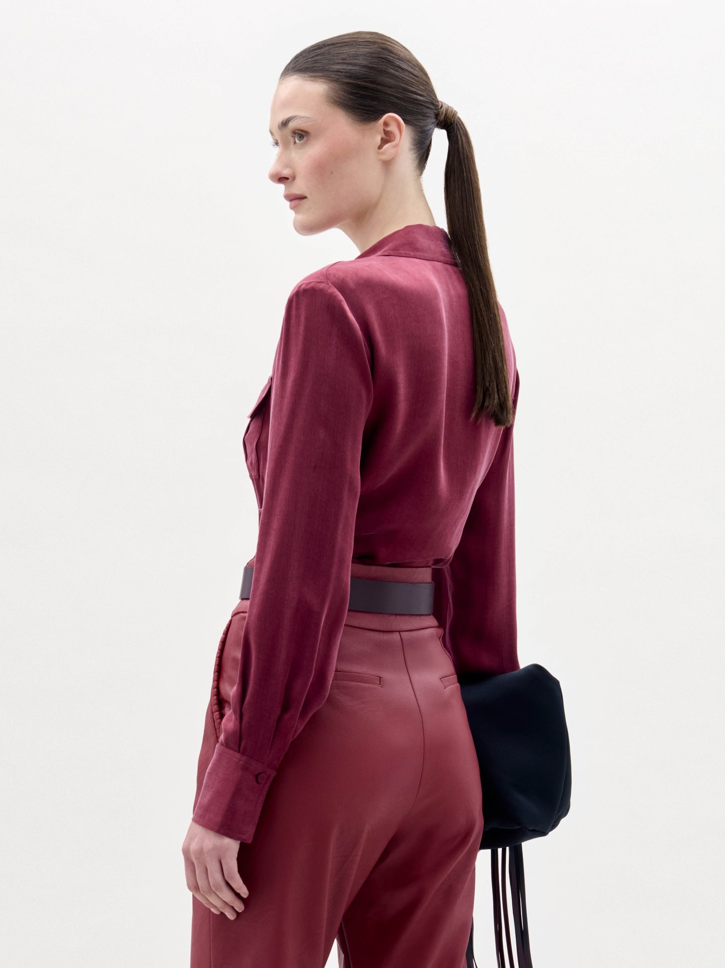 A woman stands against a plain background wearing the Caserta Blouse in deep red, matching pants, a large decorative belt, and holding a black clutch bag.