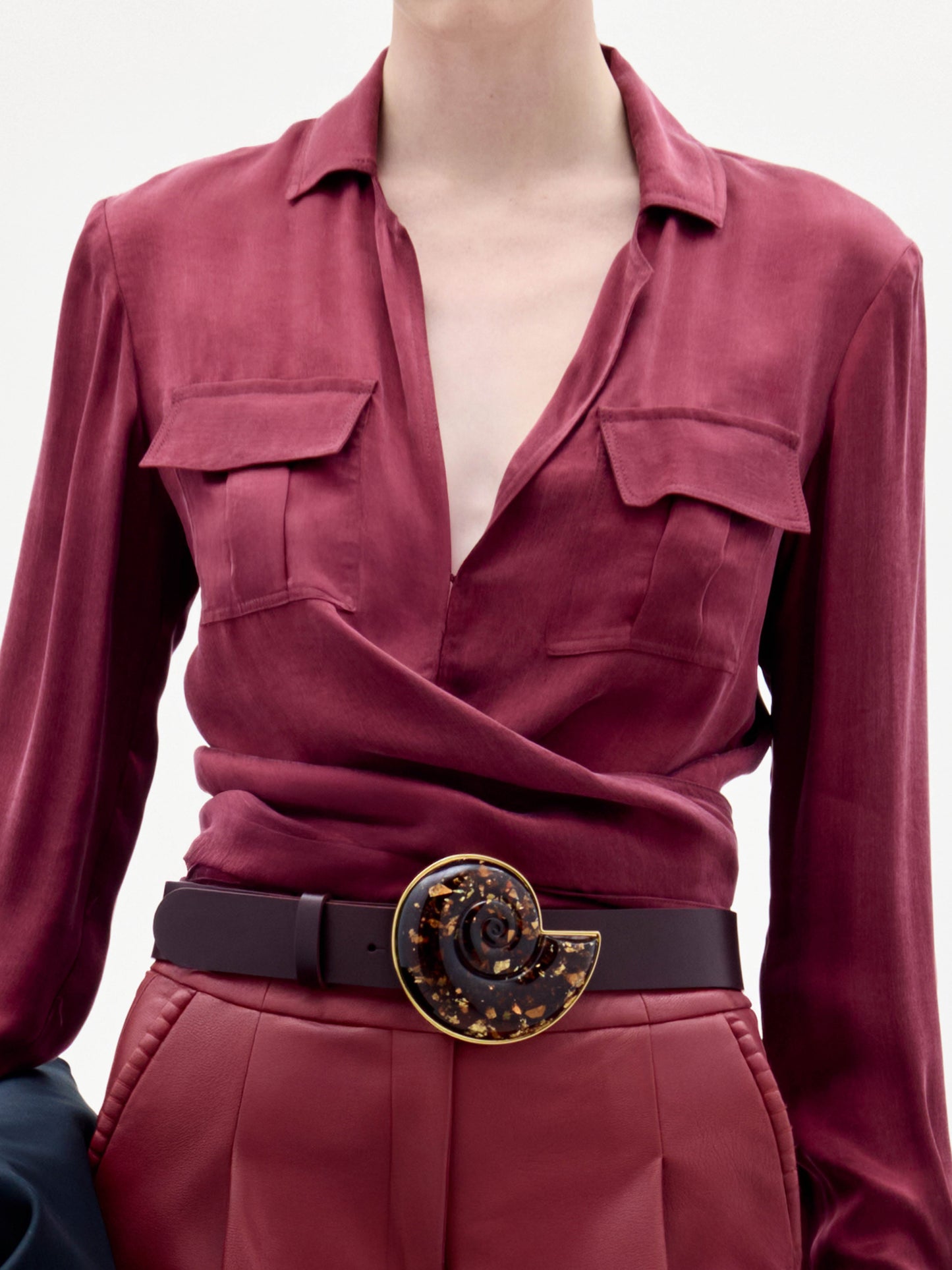 A woman stands against a plain background wearing the Caserta Blouse in deep red, matching pants, a large decorative belt, and holding a black clutch bag.