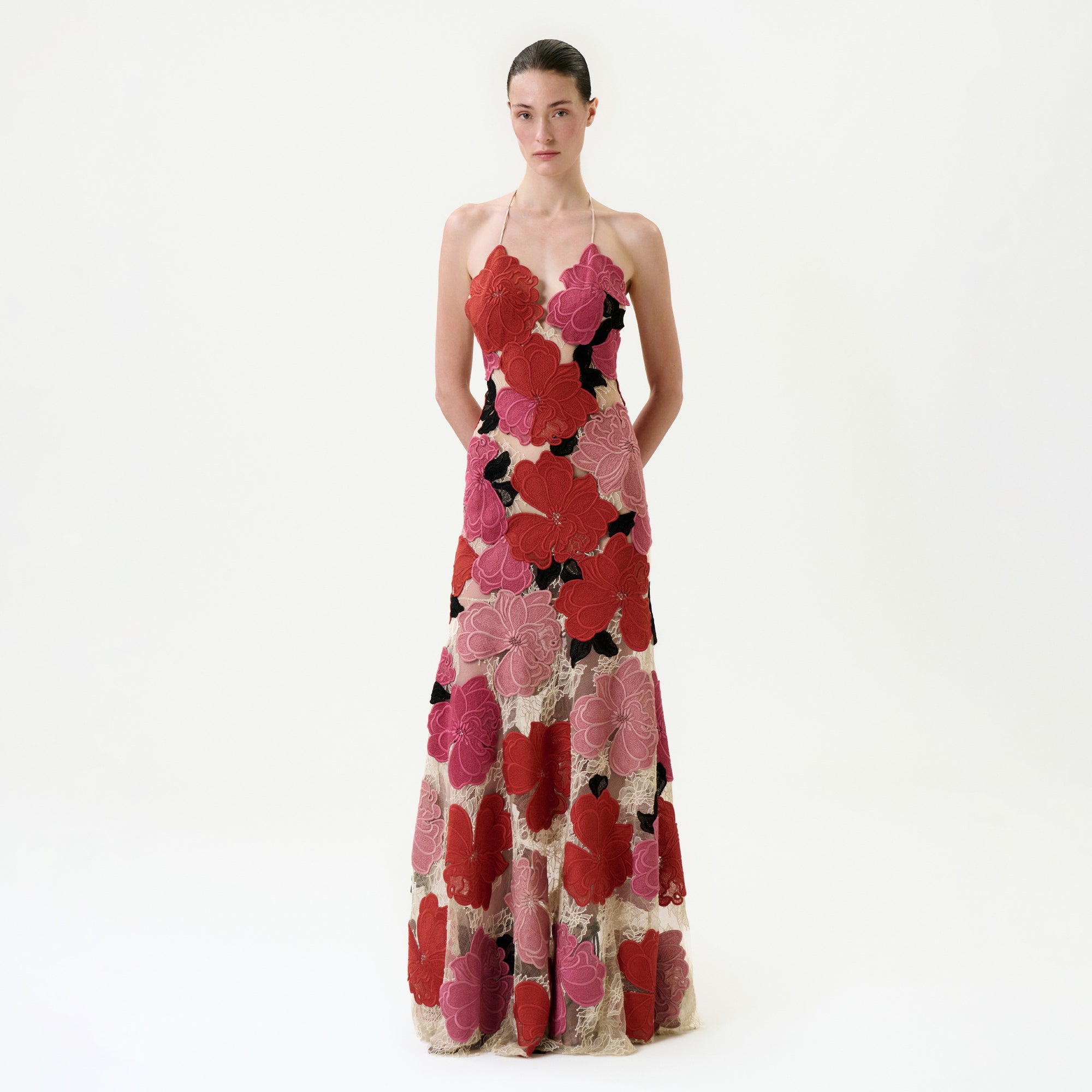 A woman with a neutral expression stands against a white background wearing the Vanessa Dress, a Colombian-made maxi dress with thin straps and bold red, pink, and black floral patterns on a beige base. Her hands are behind her back.