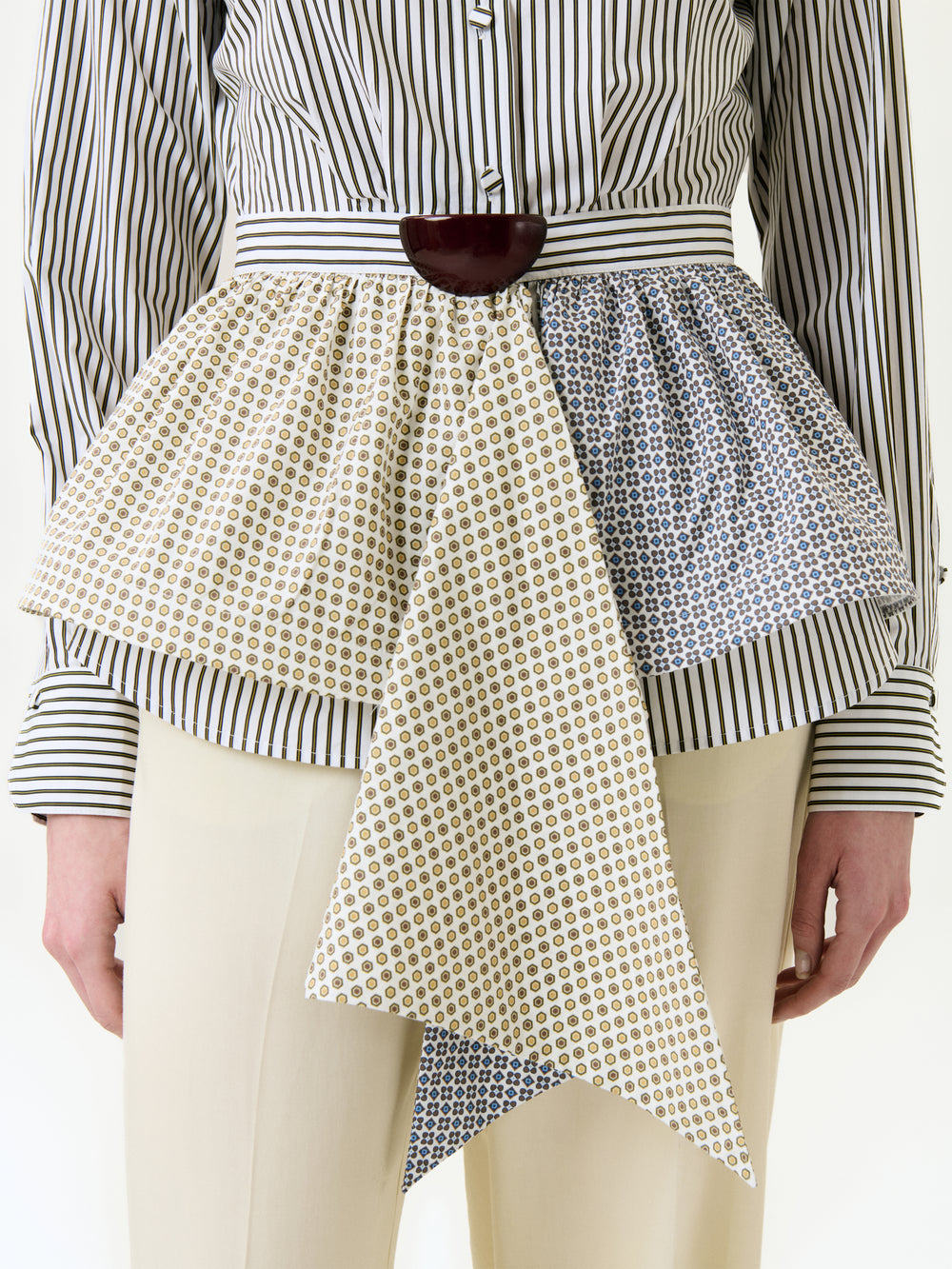 A person wears the Malina Shirt—a striped, 100% cotton button-up—paired with a wide, patterned peplum belt featuring overlapping layers and a pointed front detail, and cream-colored pants. Only the torso and upper legs are visible.