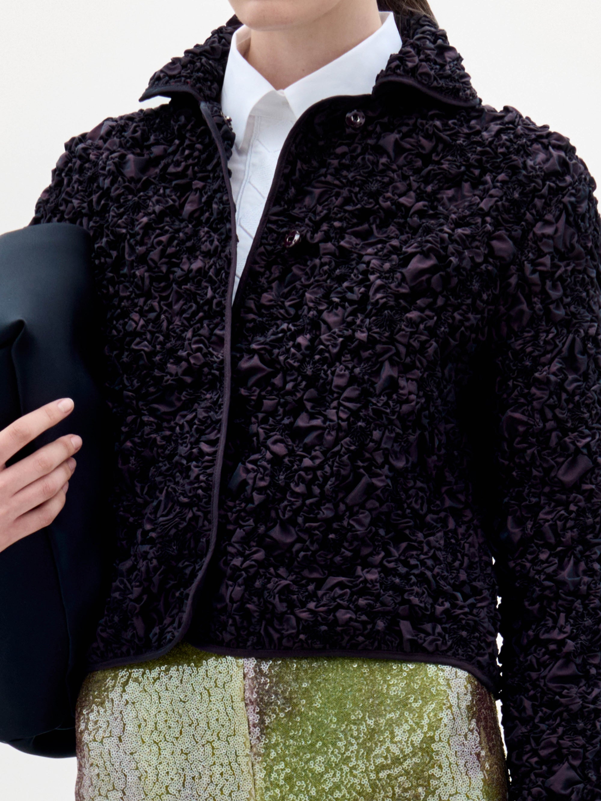 A woman stands against a plain background, wearing the textured black Jamari Jacket over a white shirt and shiny patterned pants, holding a folded black item in her left arm.