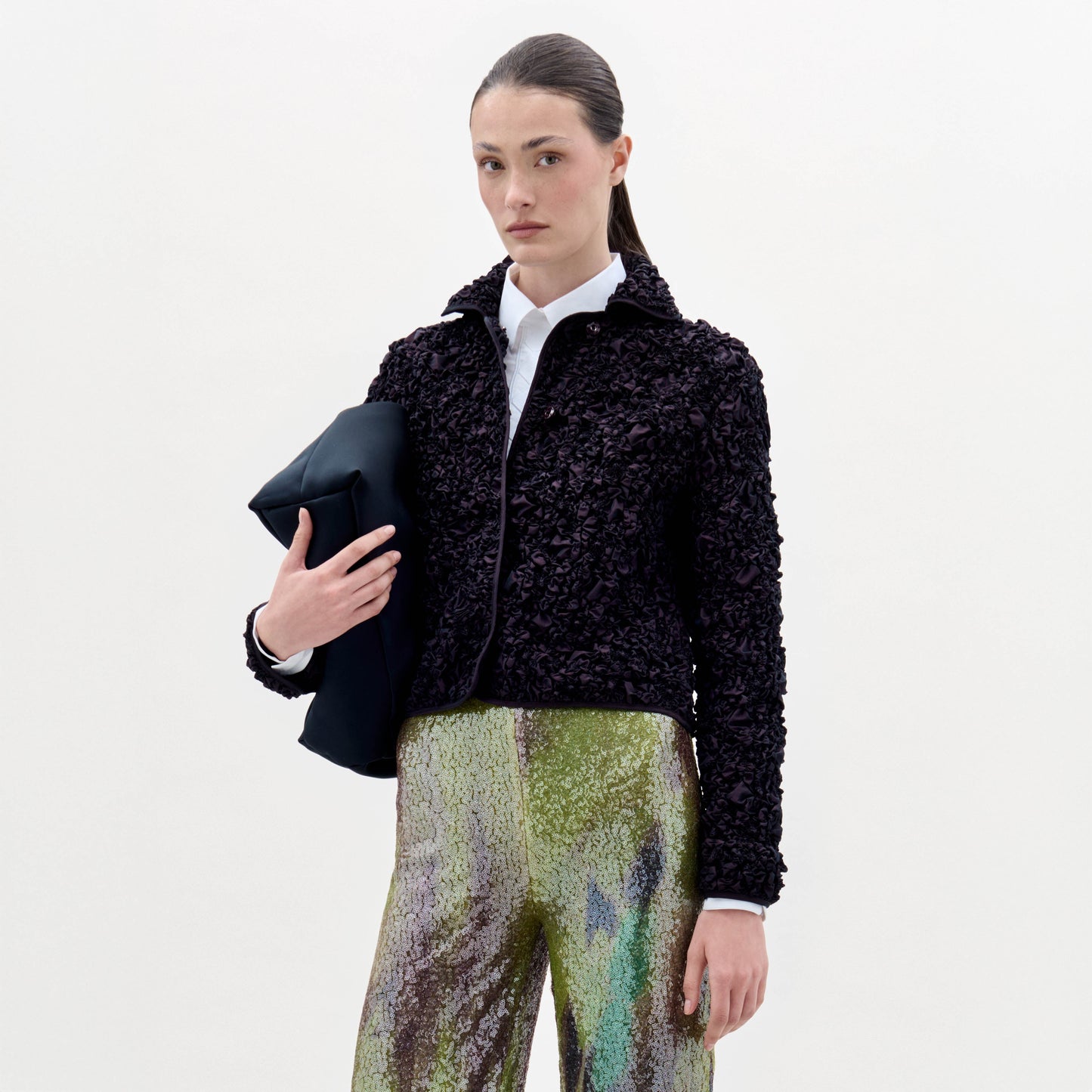 A woman stands against a plain background, wearing the textured black Jamari Jacket over a white shirt and shiny patterned pants, holding a folded black item in her left arm.