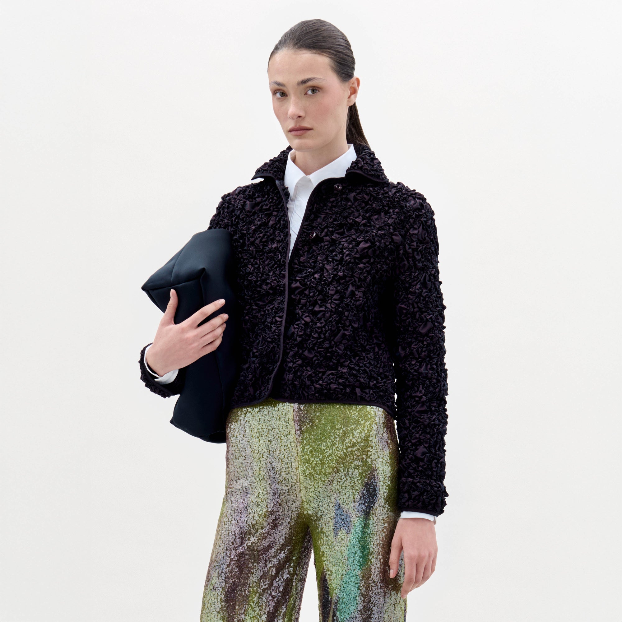 A woman stands against a plain background, wearing the textured black Jamari Jacket over a white shirt and shiny patterned pants, holding a folded black item in her left arm.