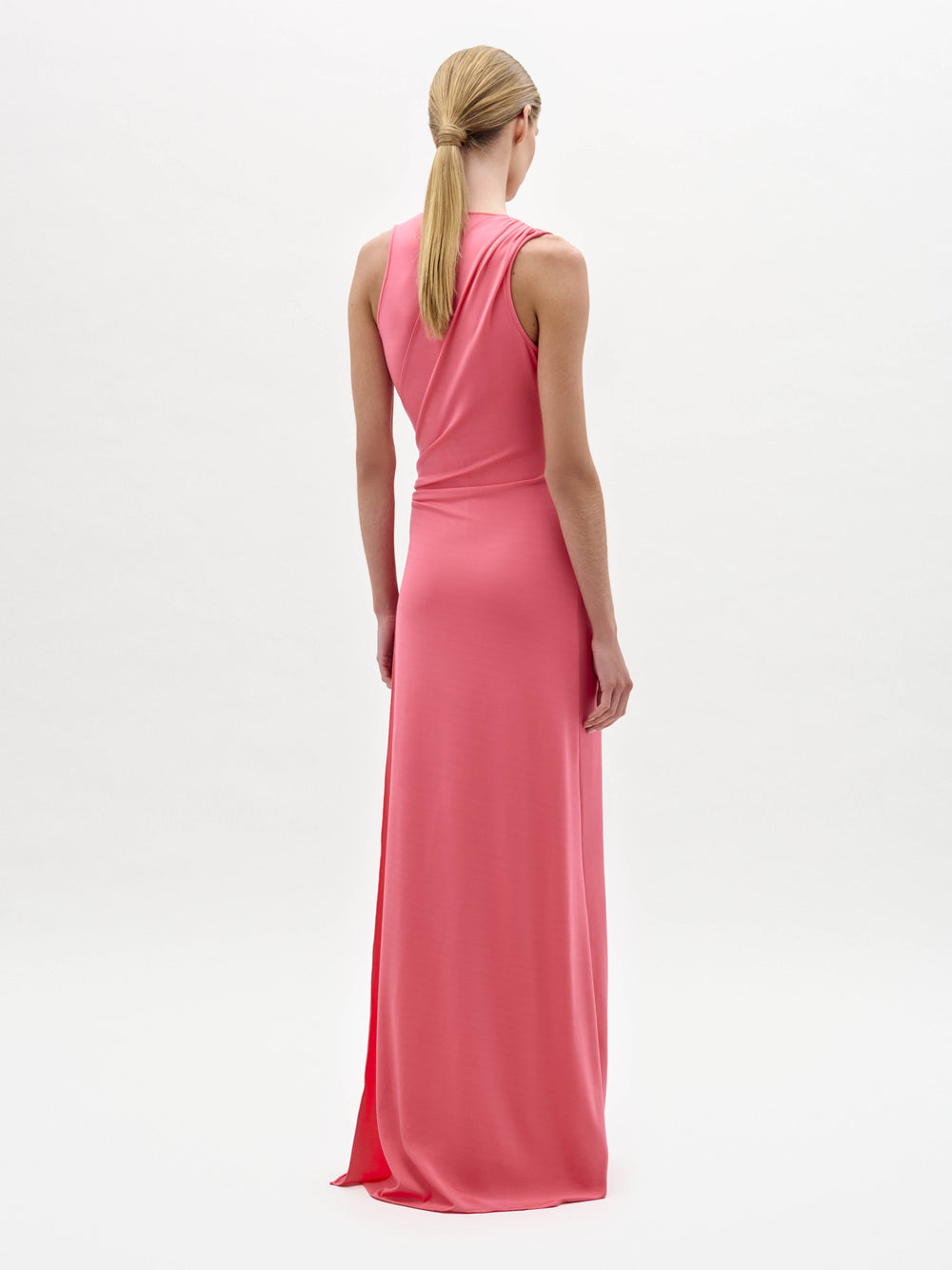 A woman stands against a white background wearing the Fadila Dress, a sleeveless, floor-length pink dress featuring gathered detailing at the waist.