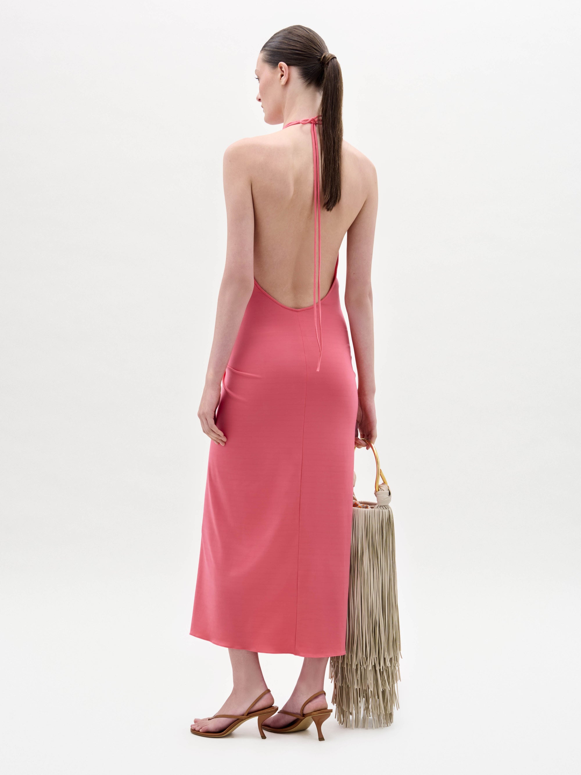 A woman stands against a plain background wearing the sleeveless pink Veracruz Dress with a gathered waist, holding a large beige fringed bag and wearing brown sandals.