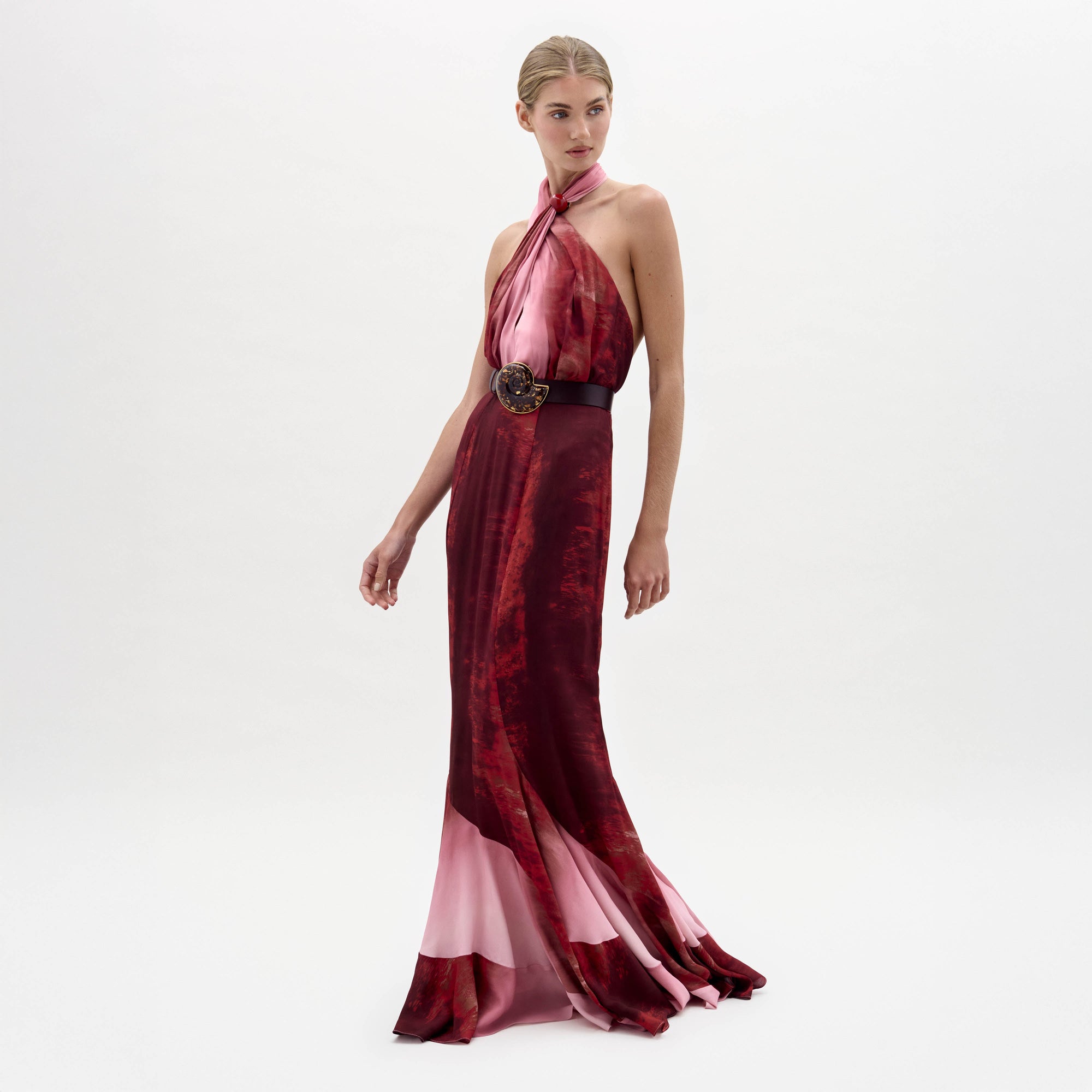 A woman stands against a plain white background wearing the sleeveless, floor-length Raisa Dress in red and pink with a wide black belt featuring a large floral buckle.