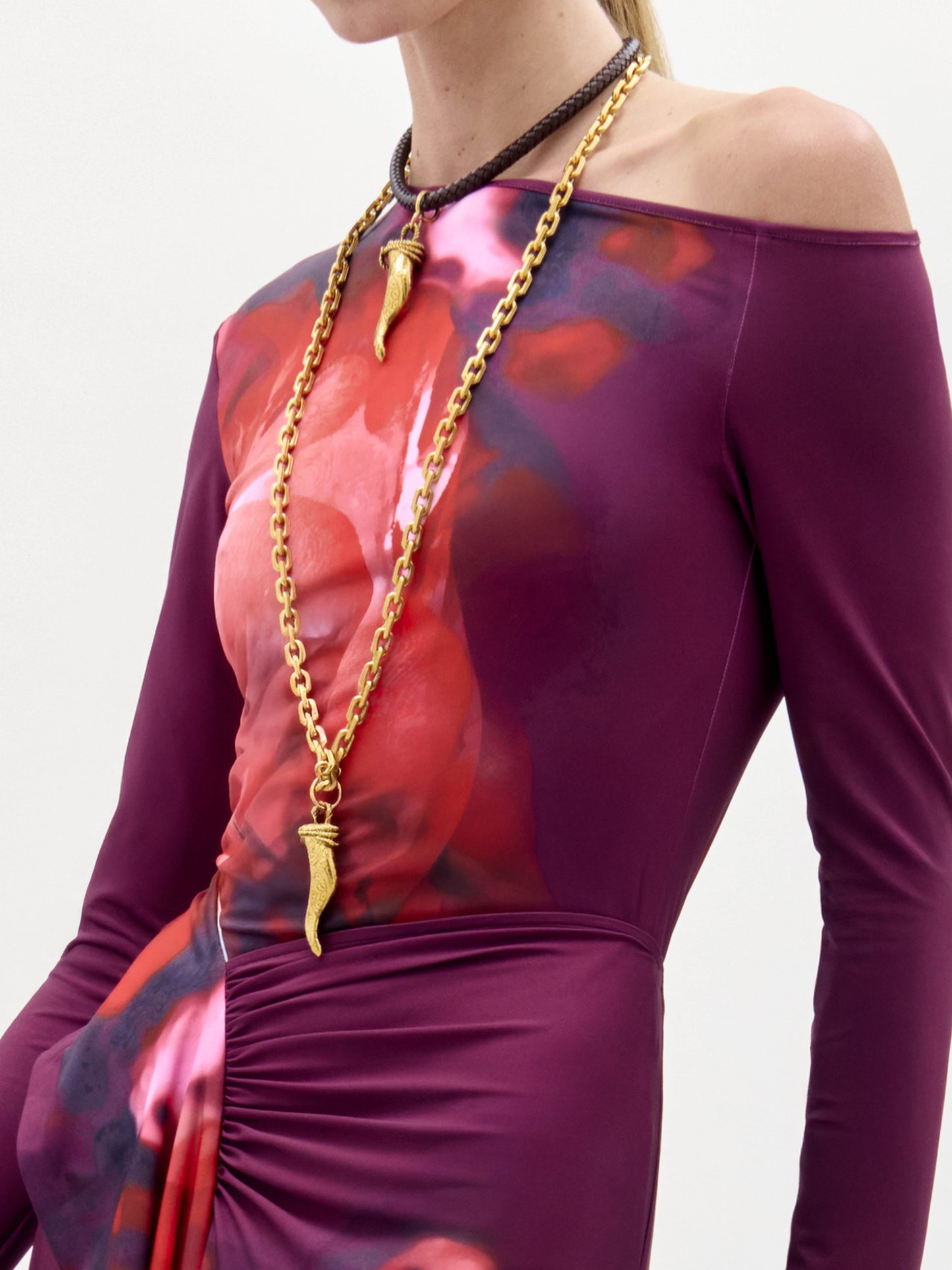 A woman wears the Sabra Blouse, a long-sleeve purple and red patterned top with an asymmetric neckline and a long gold necklace, standing against a plain white background.